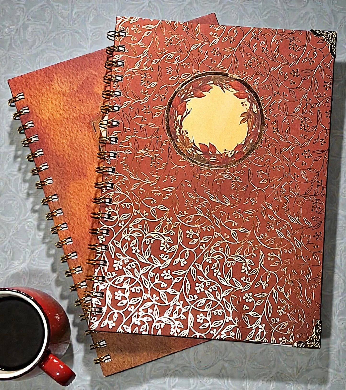 A decorative notebook with an intricate white floral pattern and a circular cutout on the front, placed on a table with a cup of coffee nearby.
