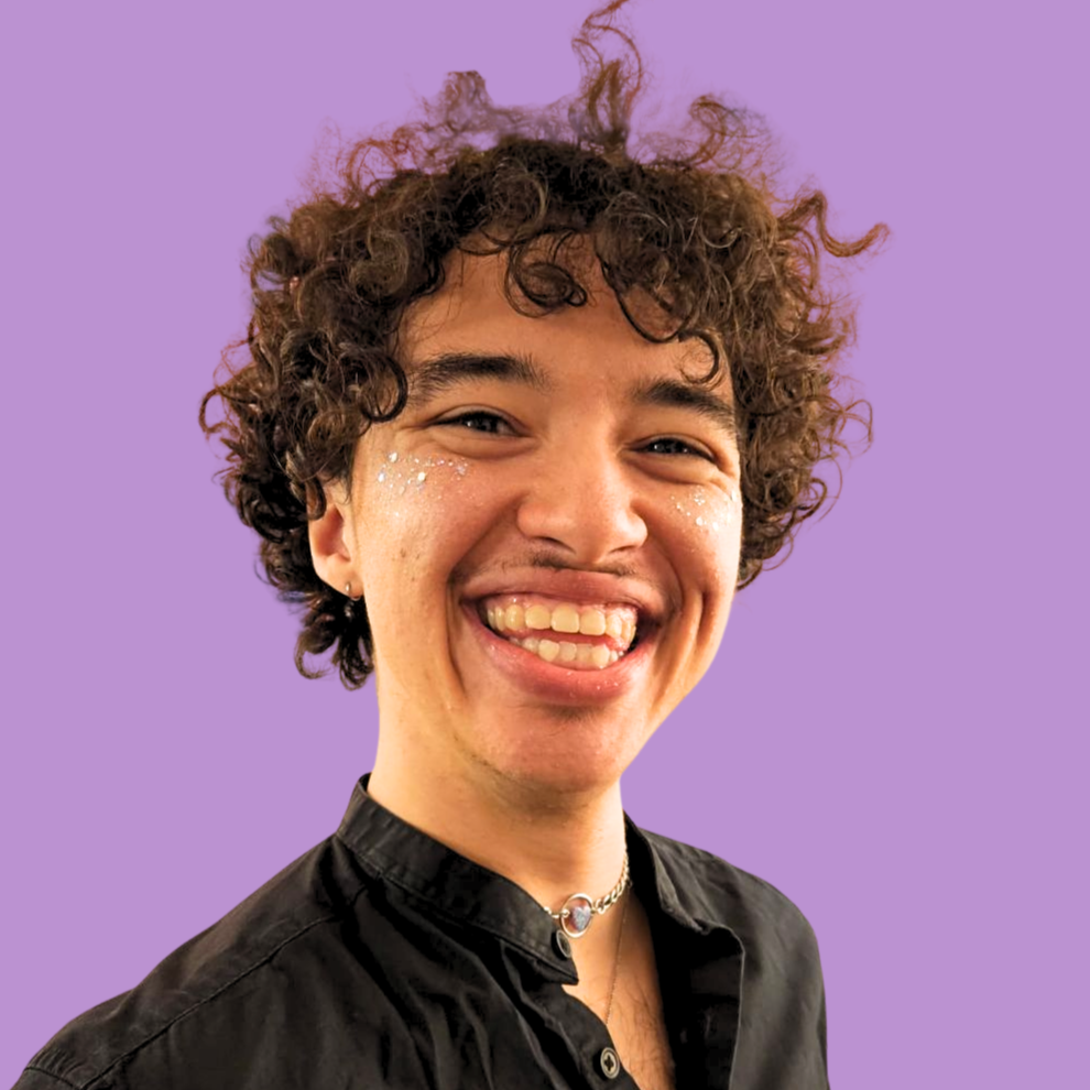 BIPOC Person with short brown curly hair, a big smile glitter dusted cheeks, sparkly heart choker necklace, and a black button up shirt. With a Purple Backdrop.