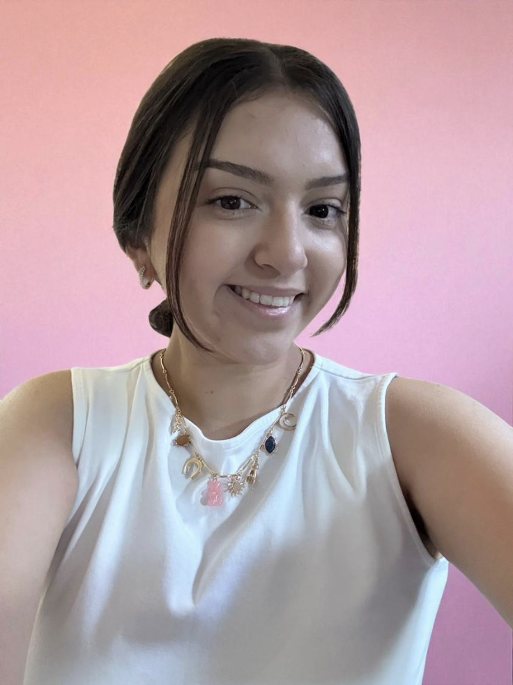 Latin-a Woman with brown hair pulled back , wearing a white scoop neck shirt and a gold and pink necklace with a pink backdrop.