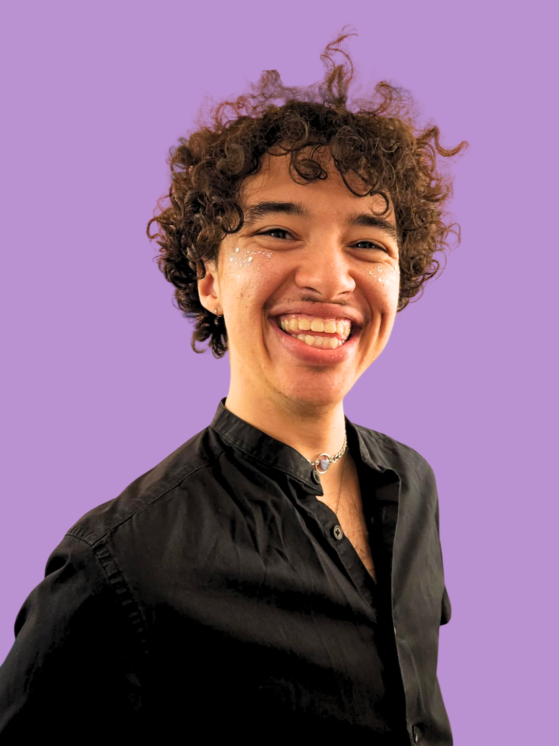 BIPOC Person with short brown curly hair, a big smile glitter dusted cheeks, sparkly heart choker necklace, and a black button up shirt. With a Purple Backdrop.