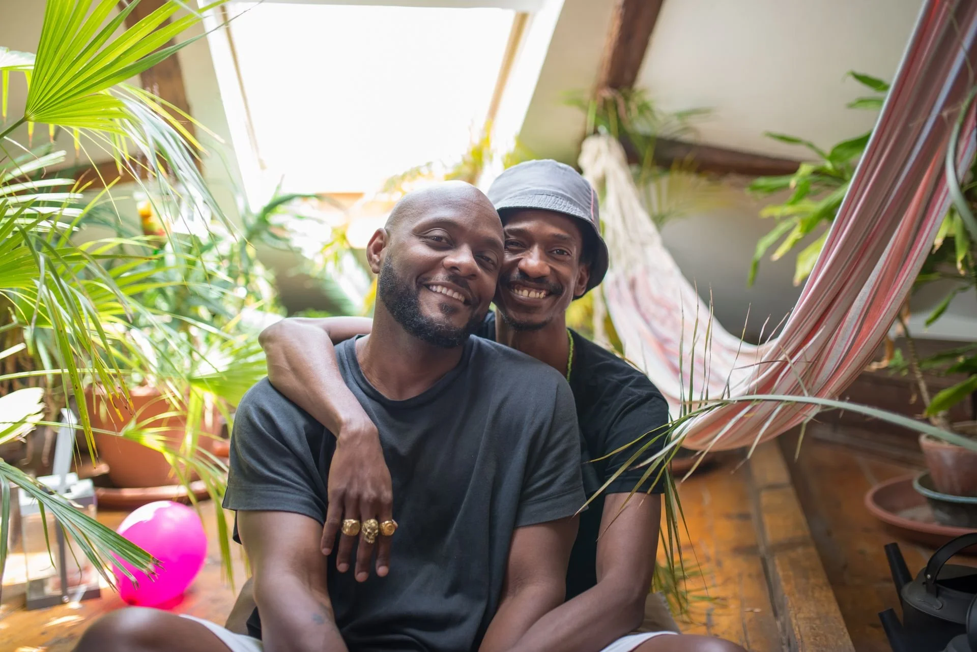 Two Black men embrace each other, facing the viewer and smiling.  The man sitting further back is wearing a bucket hat and has his arm draped around his companion. The men sit in a warmly lit room, surrounded by lush, green plants.