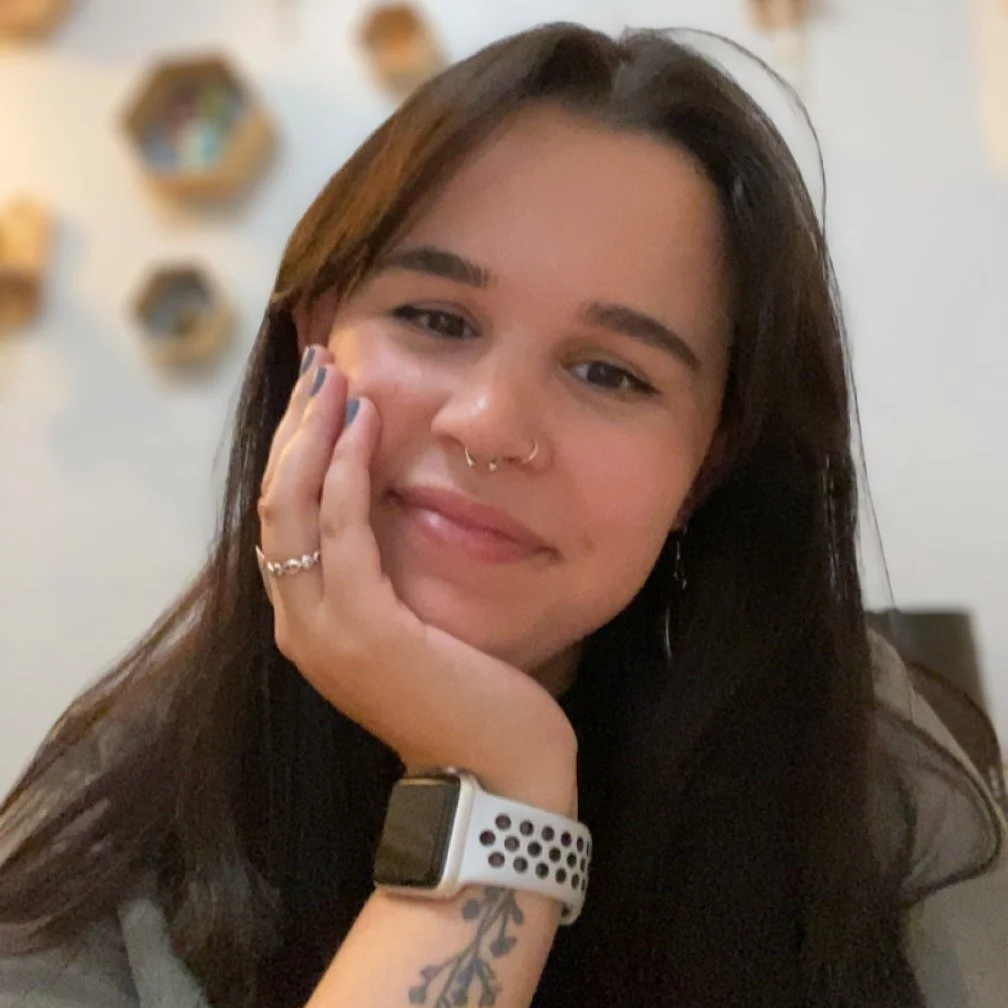 A young woman with long brown hair, wearing a watch, with a tattoo on her wrist, and a septum piercing, smiling and resting her face on her hand in a home setting with decorative wall shelves in the background.