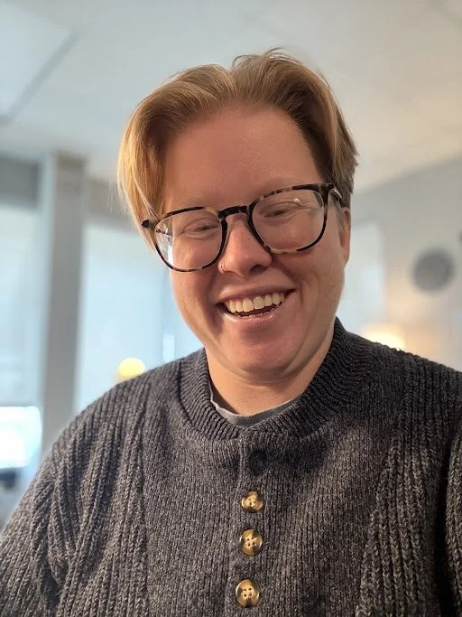 White person smiling with short blond hair, wearing large glasses and a white sweater.