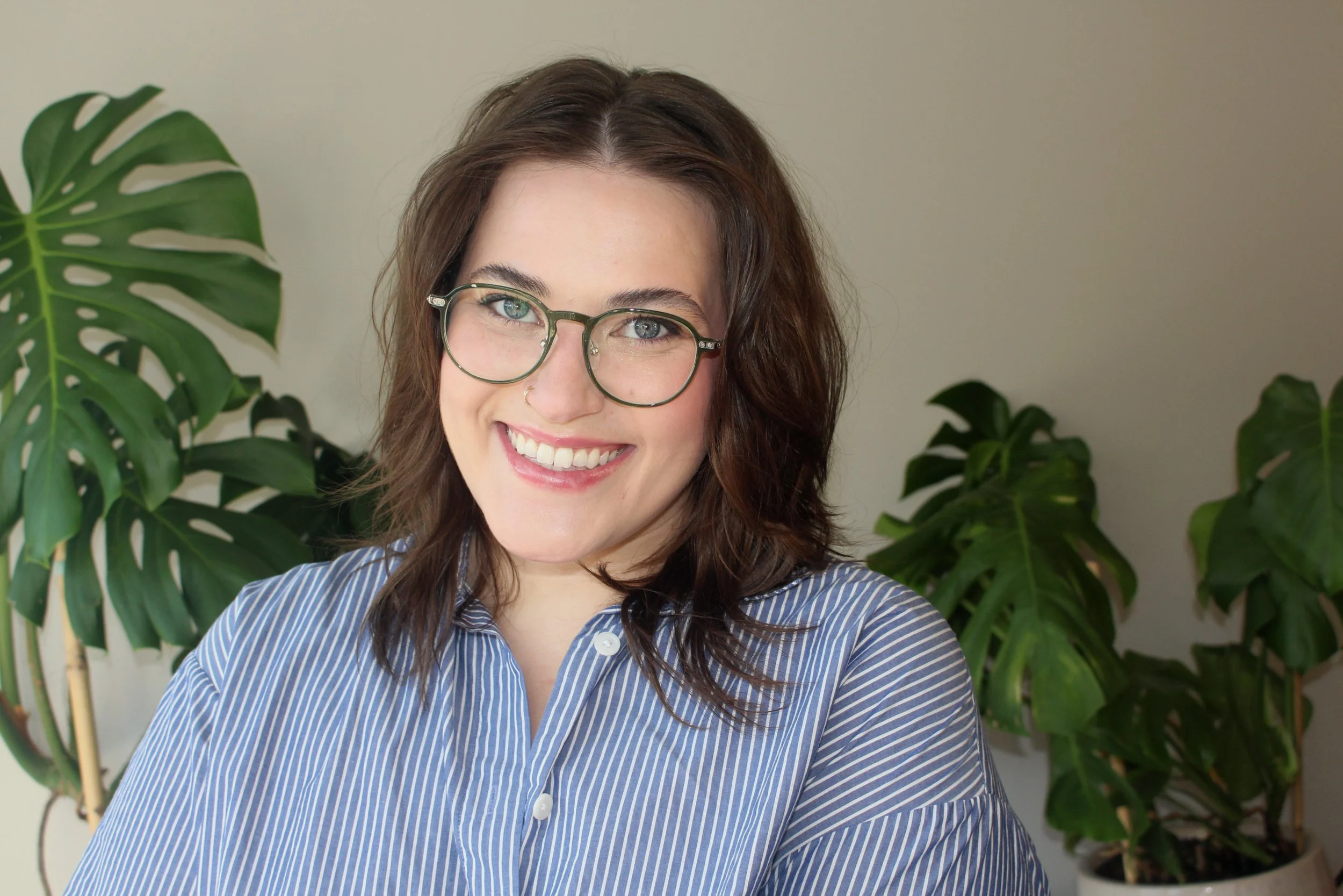 Smiling person with shoulder length brown hair, glasses, and white striped button down shirt against a beige background with tropical plants.