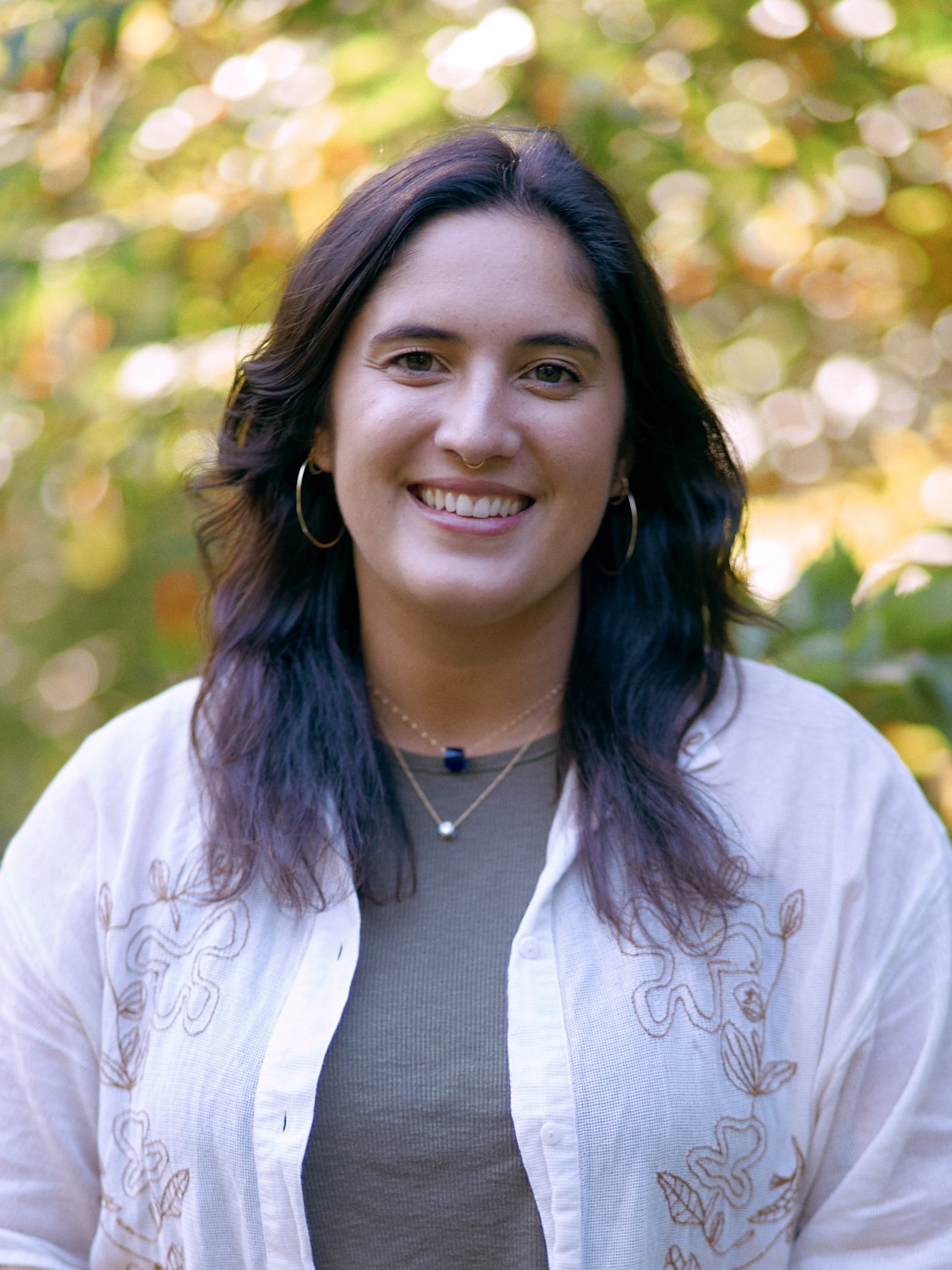 Person smiling with medium length dark wavey hair, wearing gold hoop earrings, a grey undershirt with a white collared shirt.