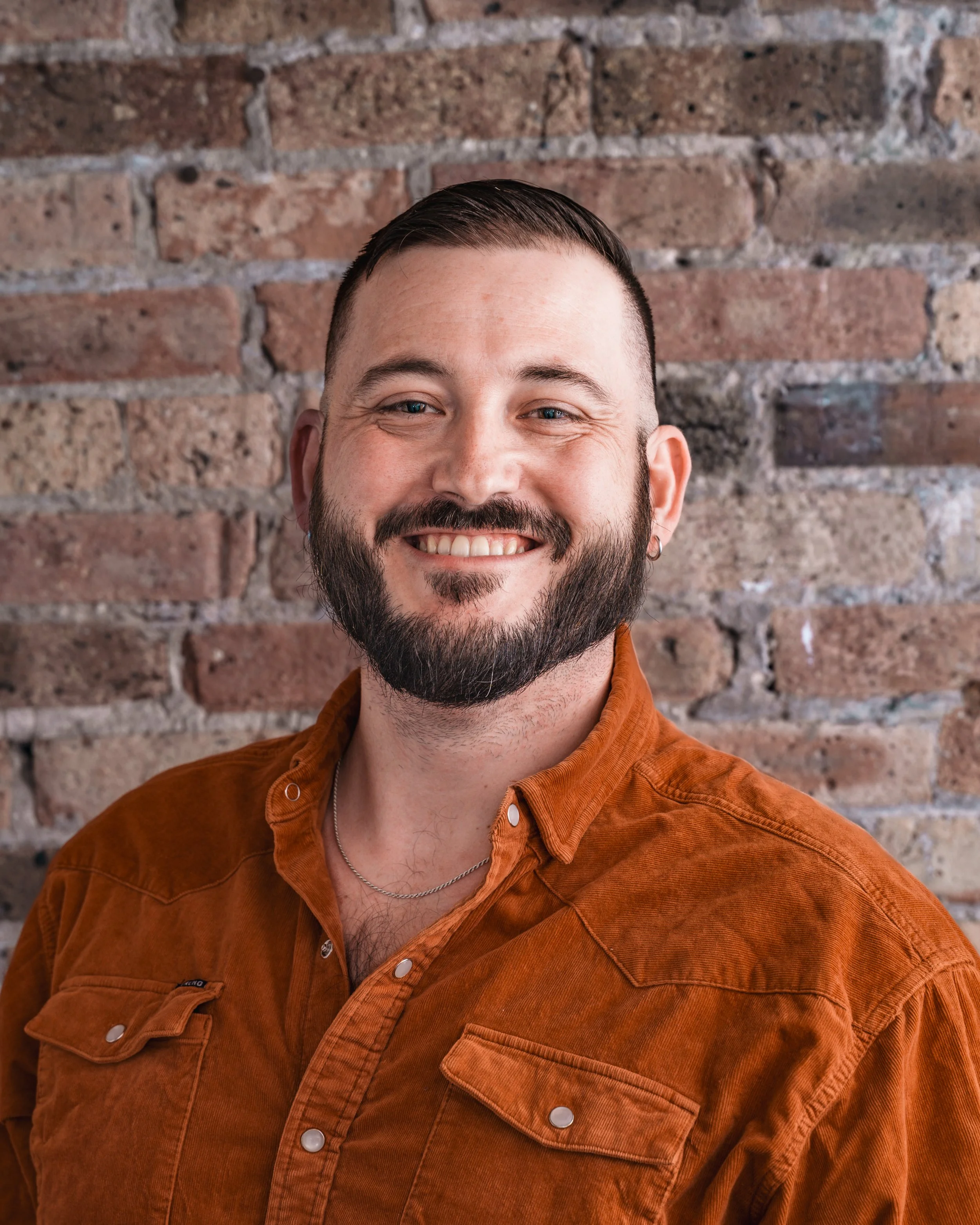 White man with short crew cut hair and a mustach, smiling, wearing a floral shirt against a gray background.