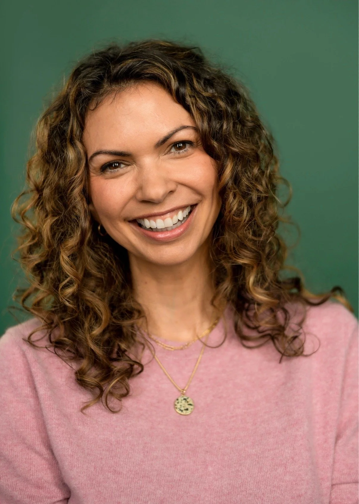 Woman with shoulder length curly hair, big smiling, wearing a pink shirt, with a dark green background