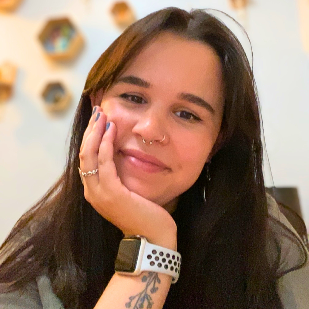 Person with a warm smile, long straight brown hair, and nose pricings, with head gently resting in palm.