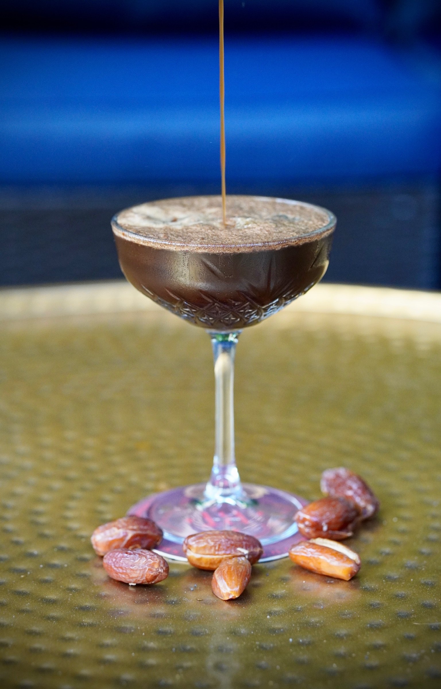 A cocktail glass with a dark drink topped with foamy froth, served on a textured yellow surface with scattered almonds around the base of the glass, and a dark blue background.