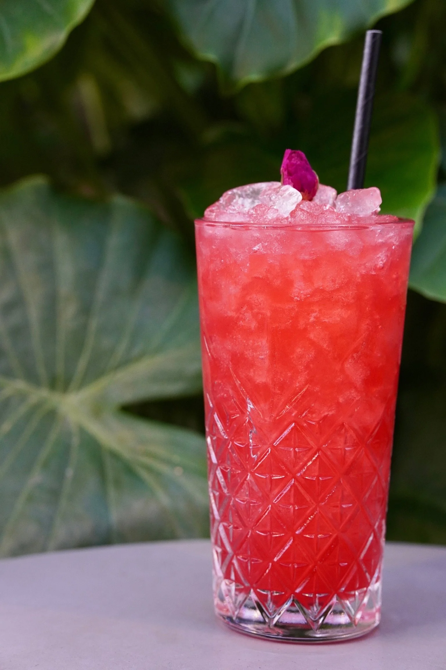 A tall glass of pink-colored cocktail with crushed ice, garnished with a small pink flower and a black straw.