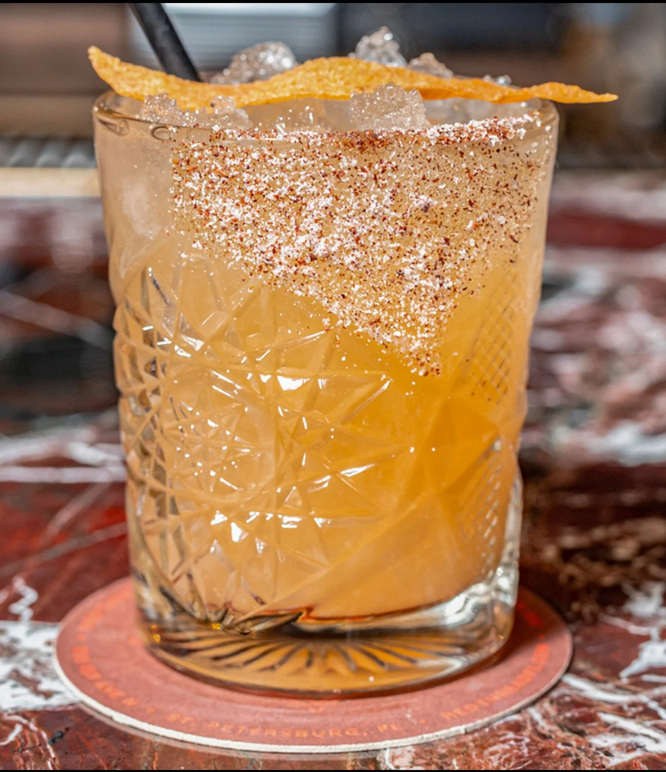 Close-up of a cocktail in a textured glass, garnished with a lemon twist, sprinkled with chili powder, with crushed ice on top.