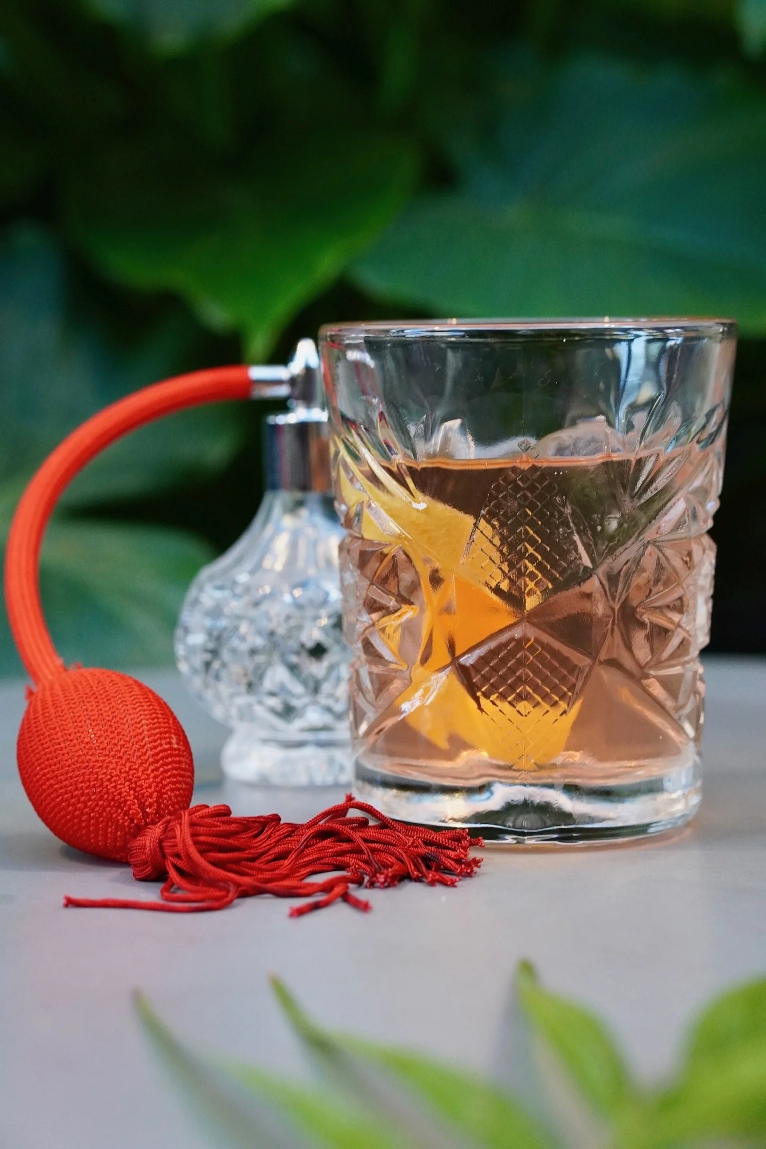 A glass of whiskey with a lemon peel garnish, accompanied by a red davidoff cigarette lighter and a clear crystal decanter, placed on a white surface with green foliage background.