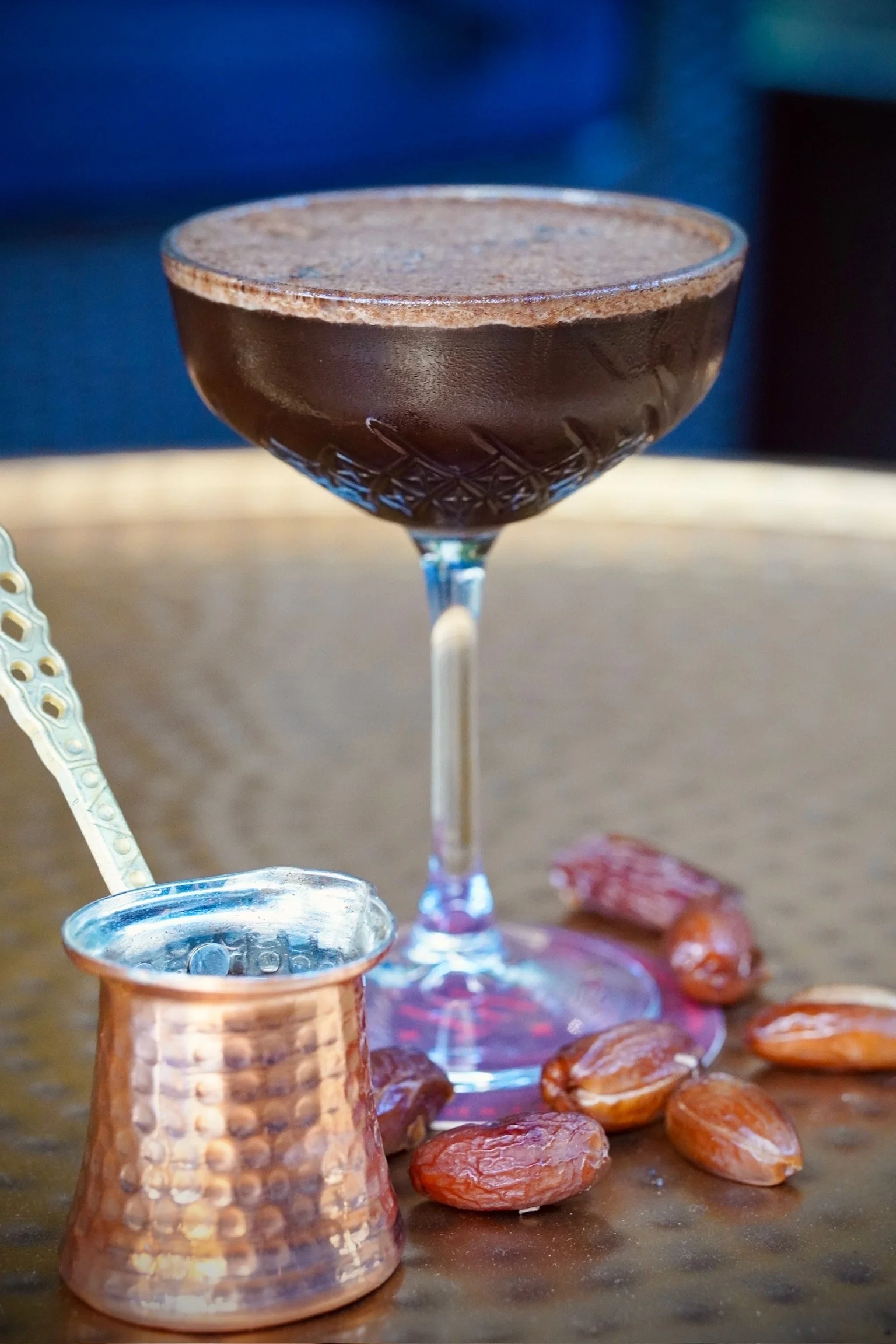 A glass of traditional Turkish coffee with a layer of foam on top, placed on a wooden surface with roasted almonds, a copper coffee pot, and a small ornate spoon nearby.