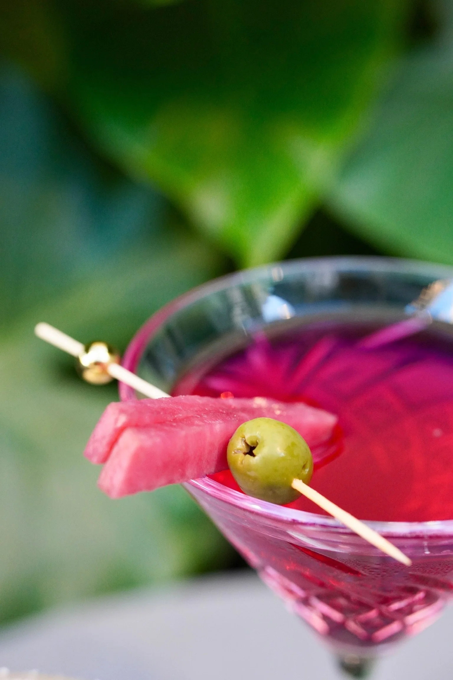 Pink cocktail glass with a slice of watermelon and a green olive on a toothpick, garnished with a pink flower, against a green blurred background.