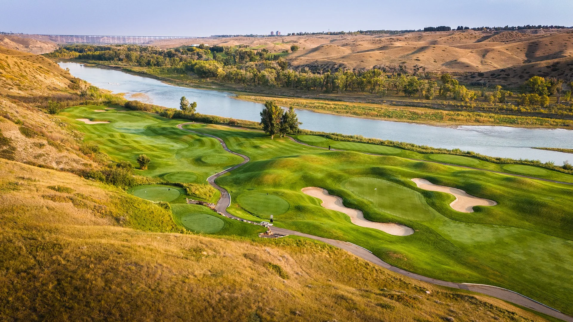 A scenic golf course located near a river, with lush green fairways, sand bunkers, and rolling hills surrounding the area.