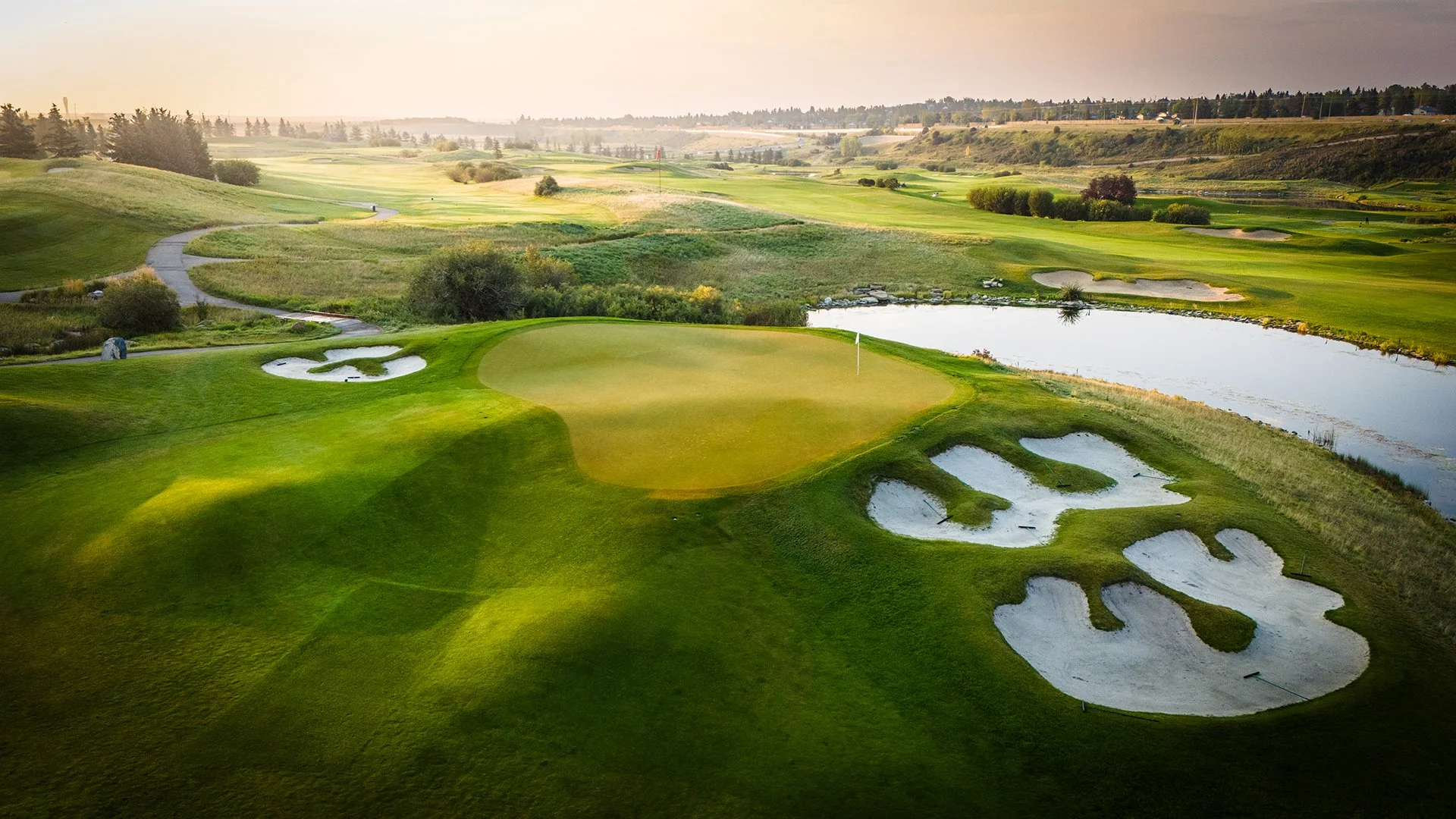 A golf course landscape at sunset with green fairways, sand bunkers, a water hazard, and a flag on the green.