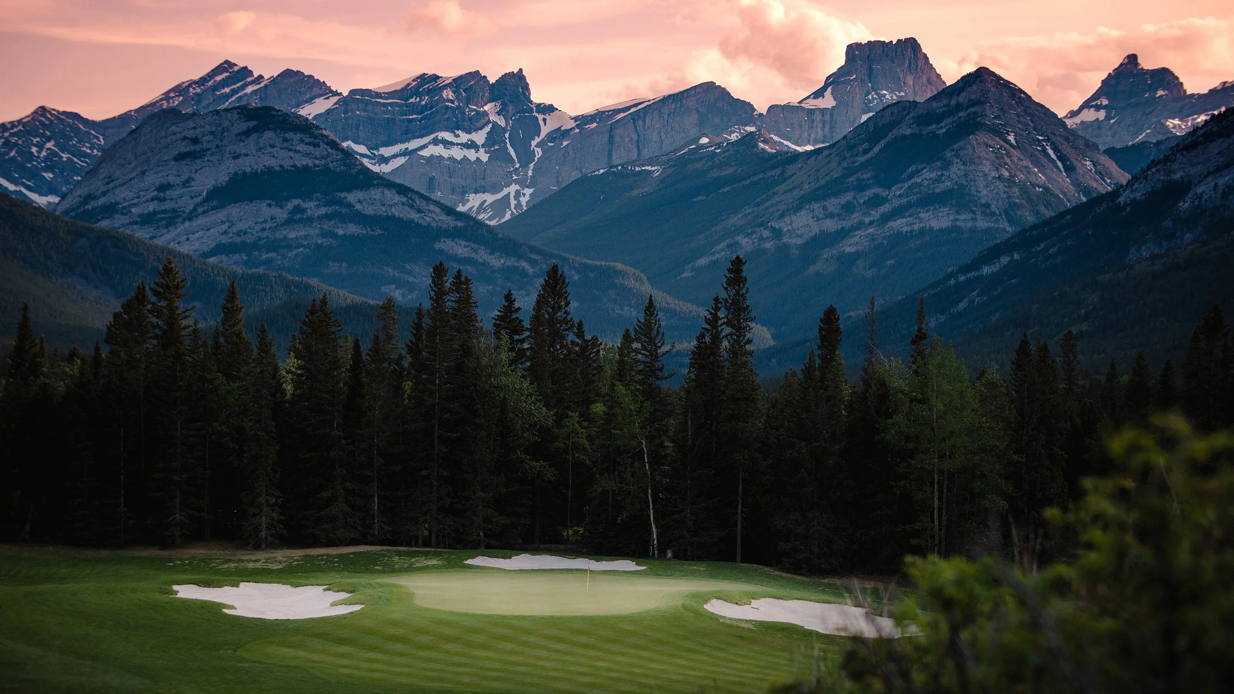 A golf course with sand bunkers and a flag on the green, surrounded by tall evergreen trees, with a backdrop of rugged snow-capped mountains under a pink and orange sky at sunset.