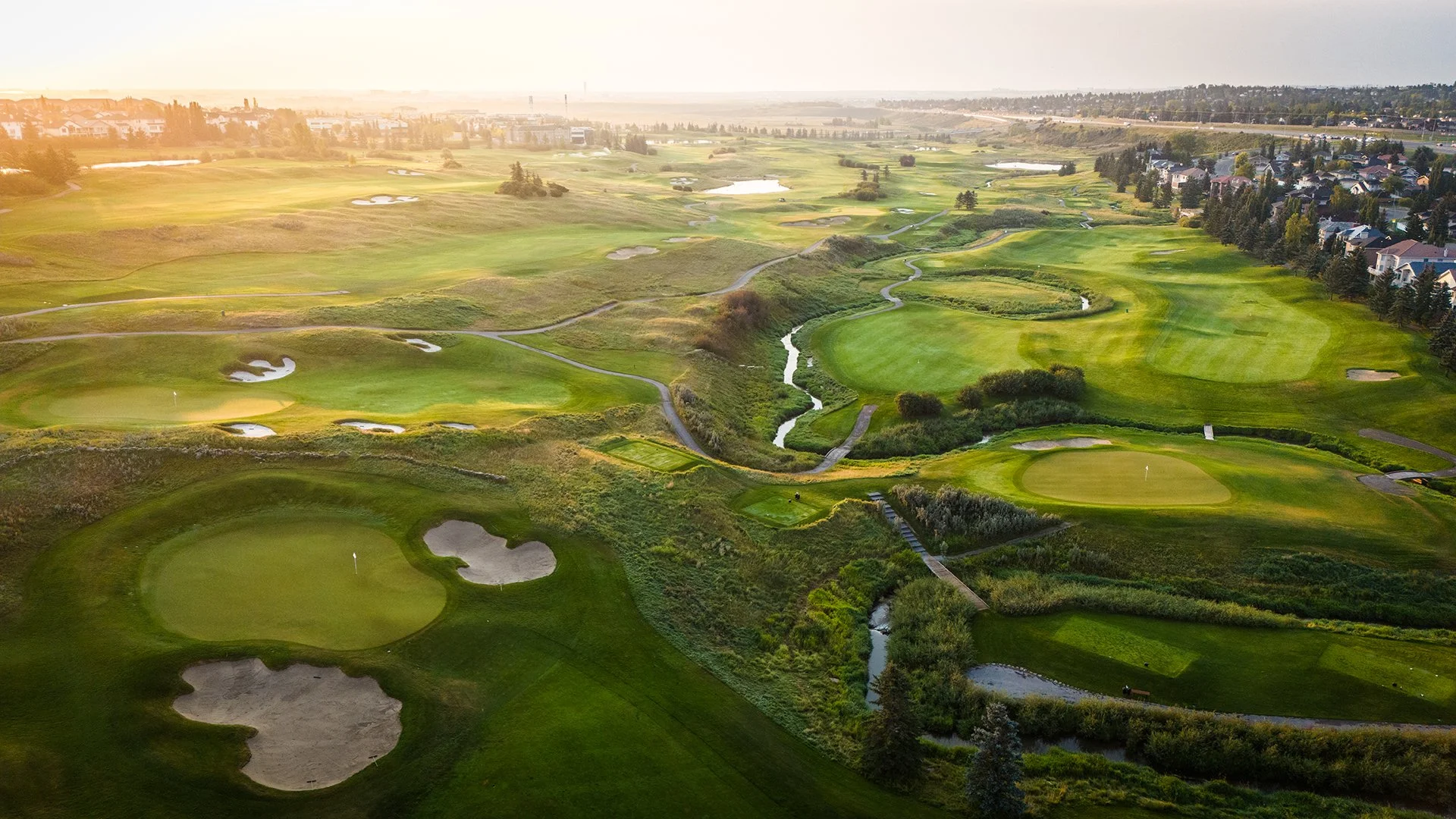 Aerial view of a golf course at sunset with green fairways, sand traps, and water hazards, surrounded by residential neighborhoods and trees.