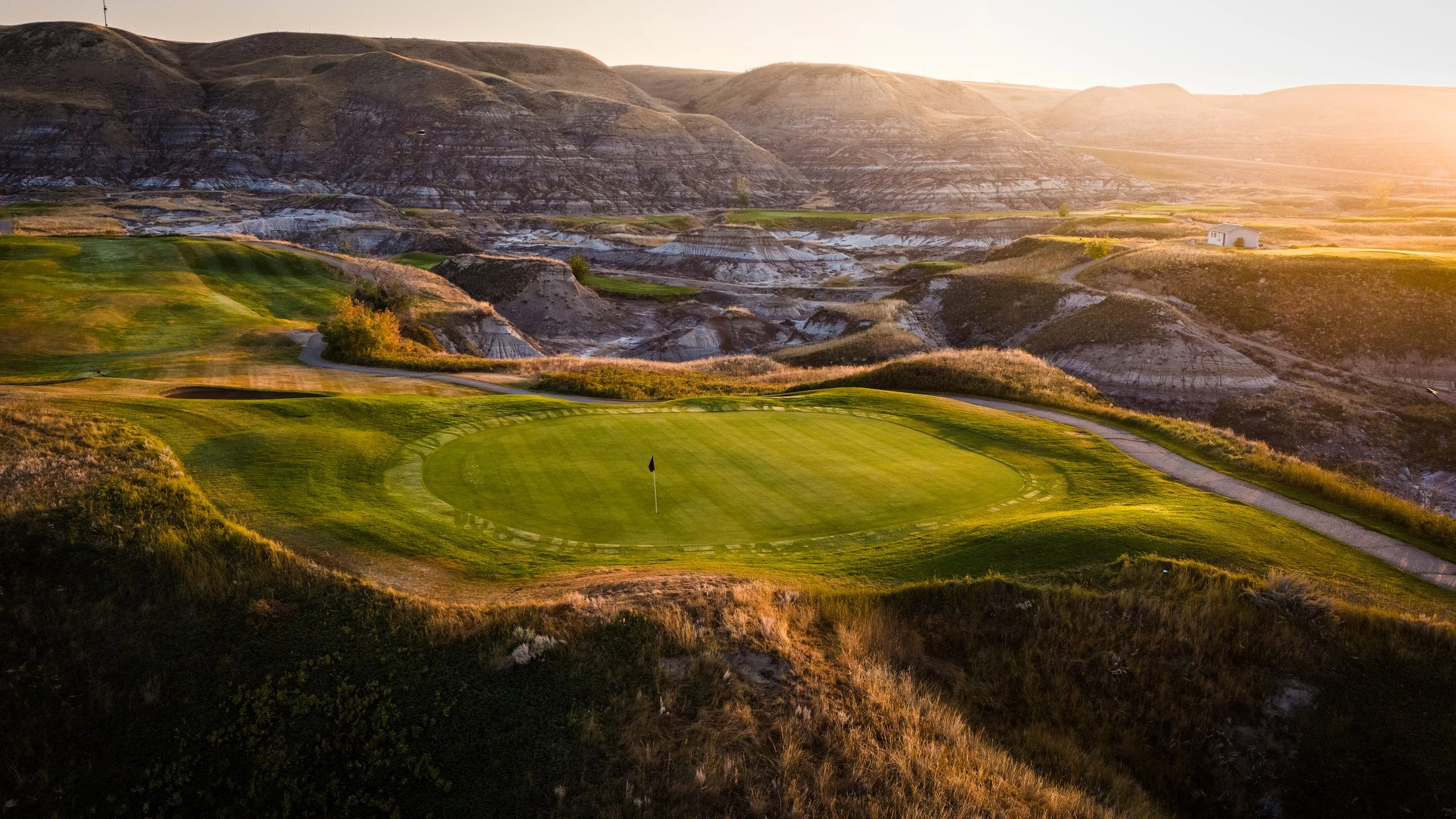 A scenic golf course with a green surrounded by sand bunkers and a flagstick, set against rugged hills and a setting sun in the background.