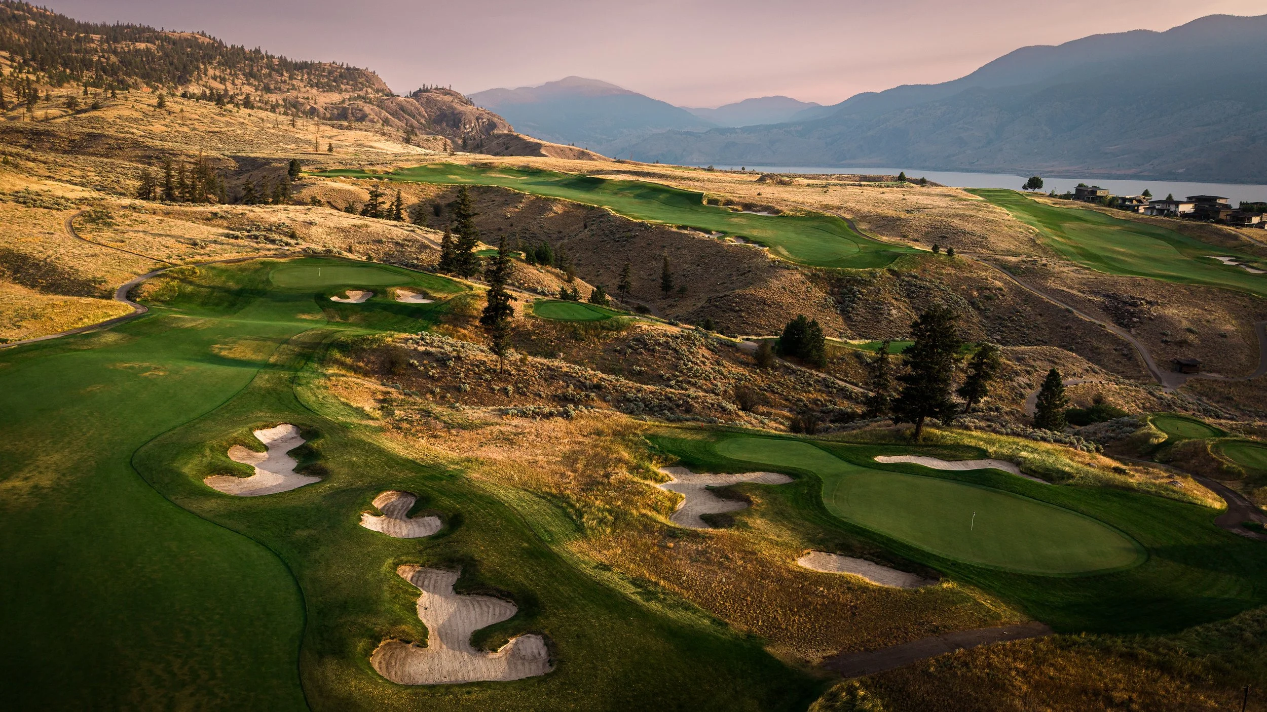A scenic view of a golf course set in a hilly landscape with mountains in the background and a lake, featuring green fairways, sand bunkers, and scattered trees.