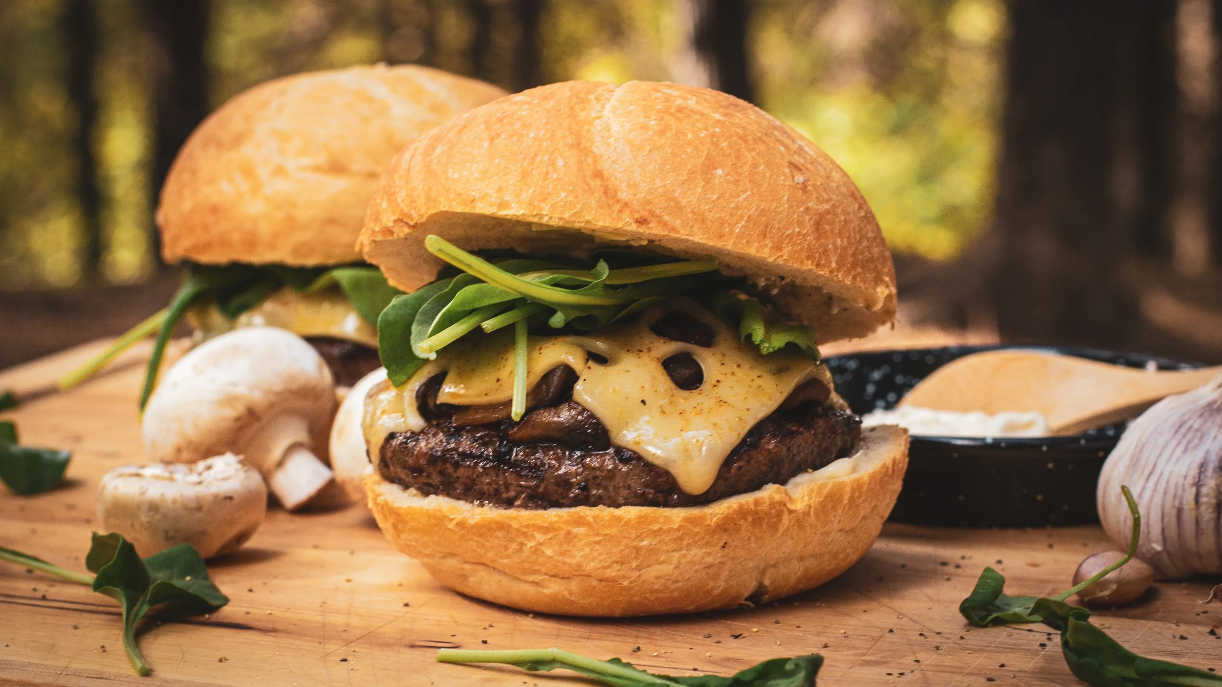 Close-up of a cheeseburger with mushrooms, greens, cheese, and a beef patty on a wooden surface, surrounded by mushrooms and garlic, with a blurred outdoor background.