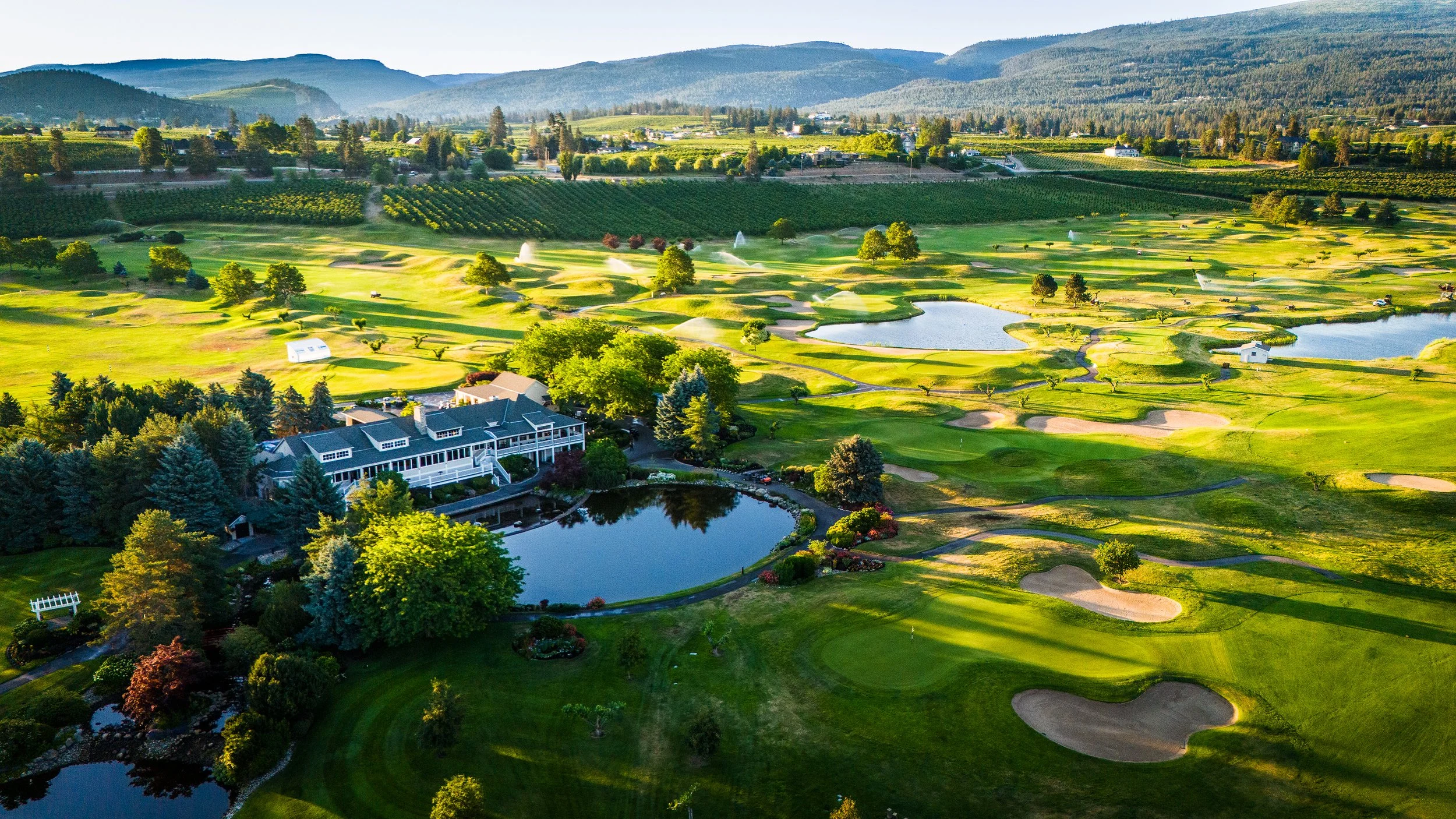 A scenic aerial view of a golf course surrounded by green hills and mountains, with water features, trees, and a building near the foreground.
