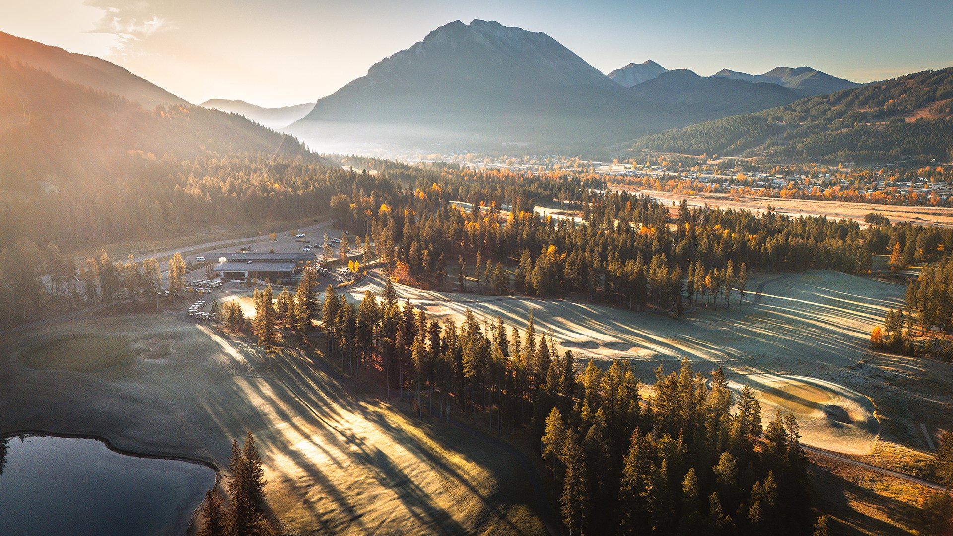 Aerial view of a golf course, mountains, and a town during sunrise, with long shadows cast by trees.