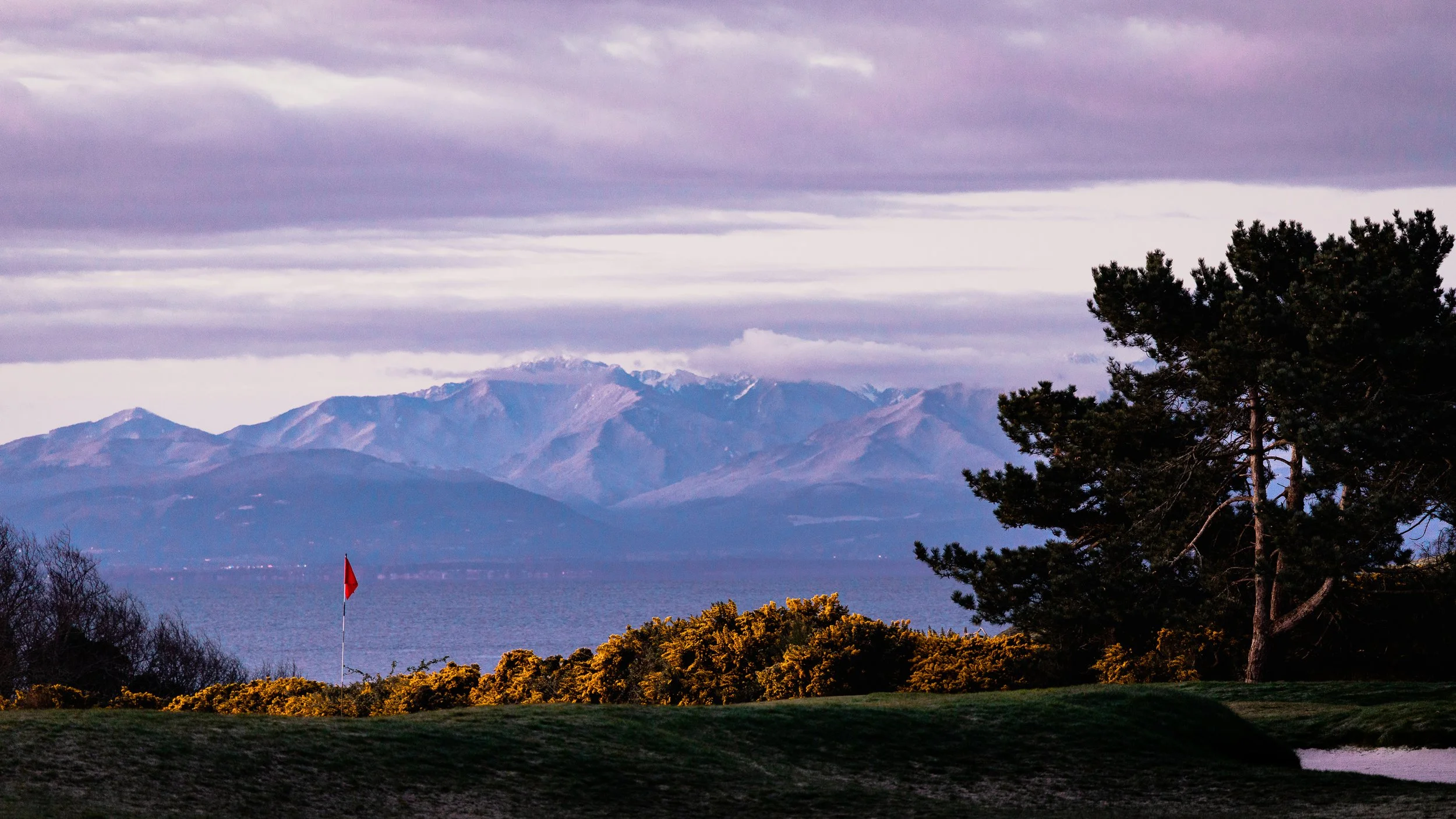 A scenic view of a lake with a red flag in the foreground, surrounded by trees, with mountains in the background under a cloudy sky.