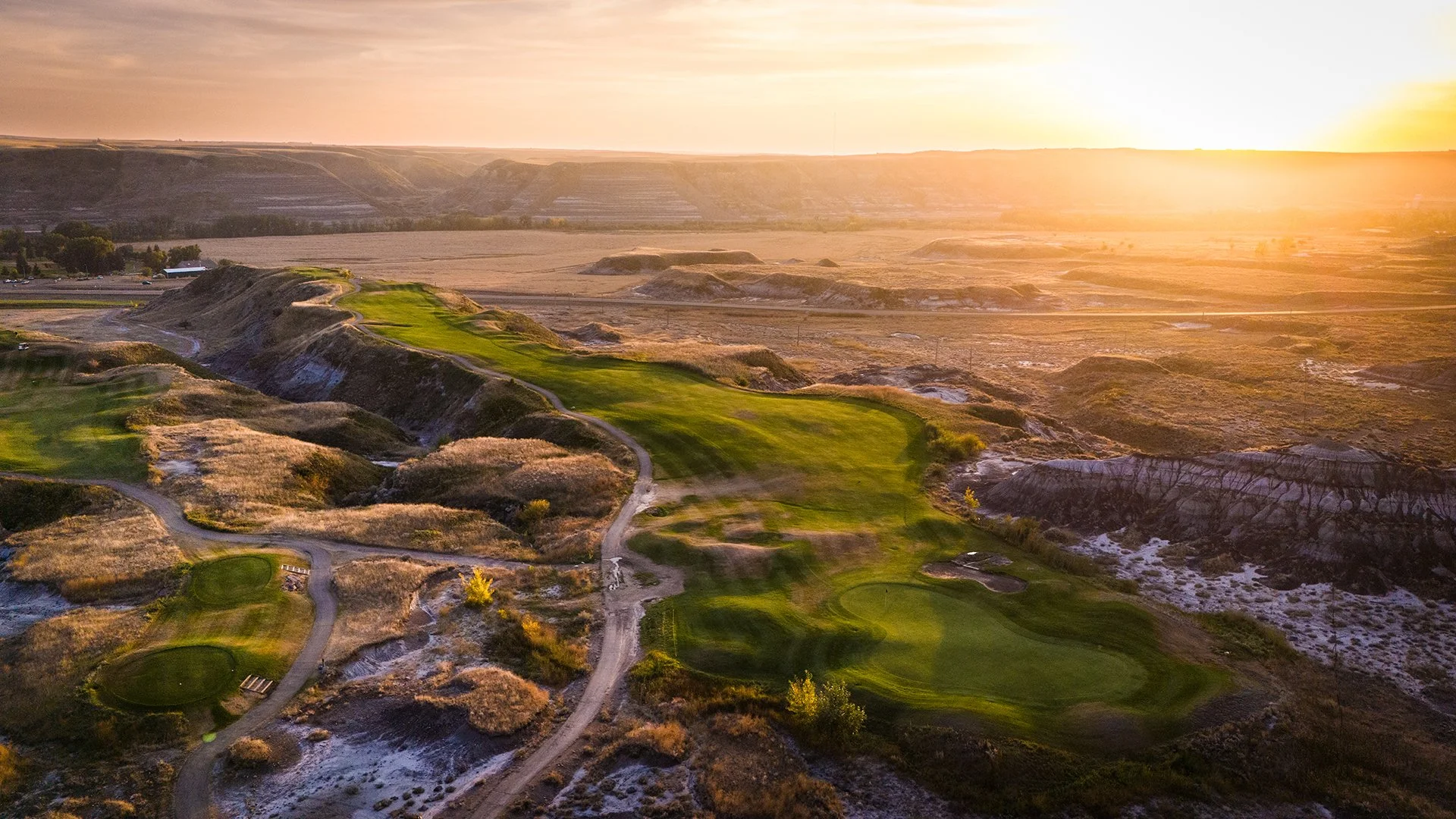 Aerial view of a golf course on a hillside at sunset, with green fairways and barren land around it.
