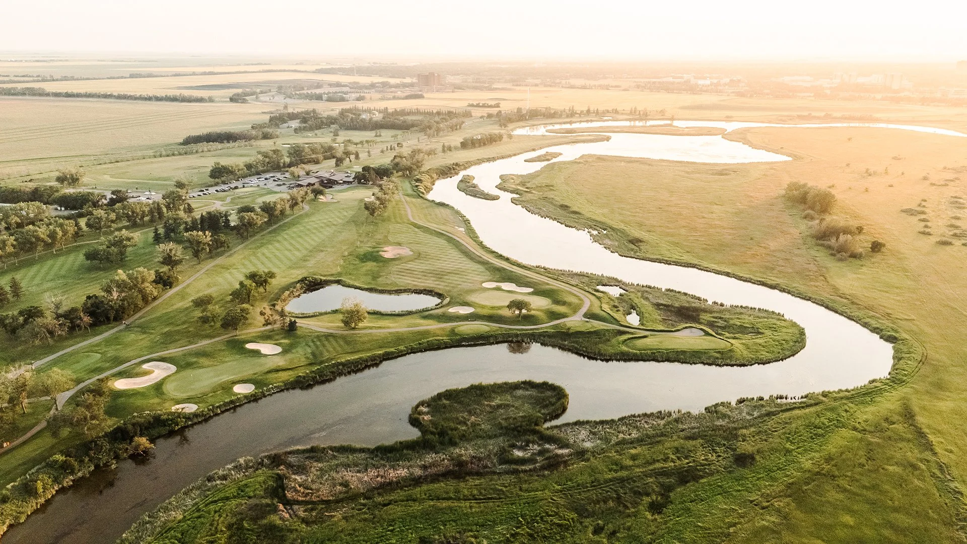 Aerial view of a golf course with multiple water hazards, sand traps, and fairways, surrounded by trees and fields, with a distant city skyline.