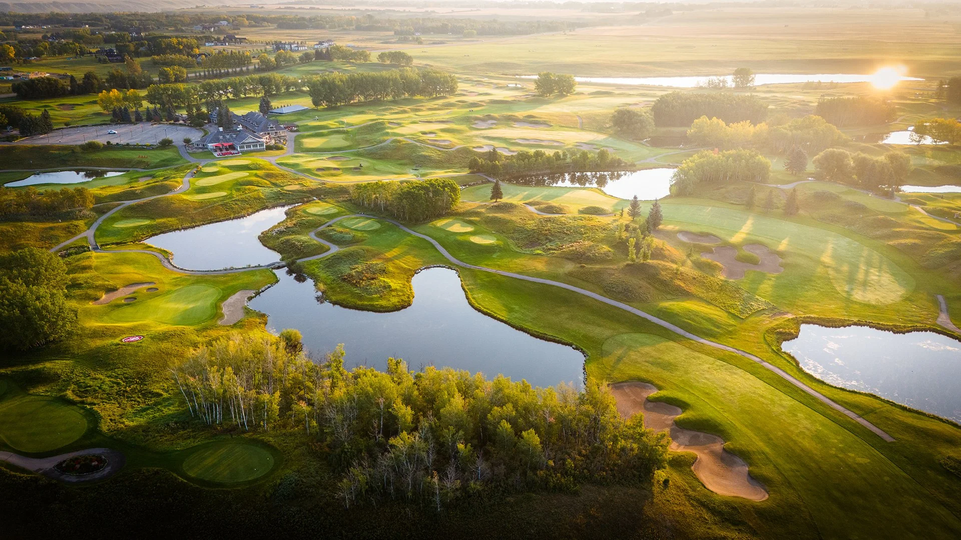 An aerial view of a golf course with several water hazards, sand traps, and green fairways surrounded by trees, with a clubhouse visible in the distance during sunset.