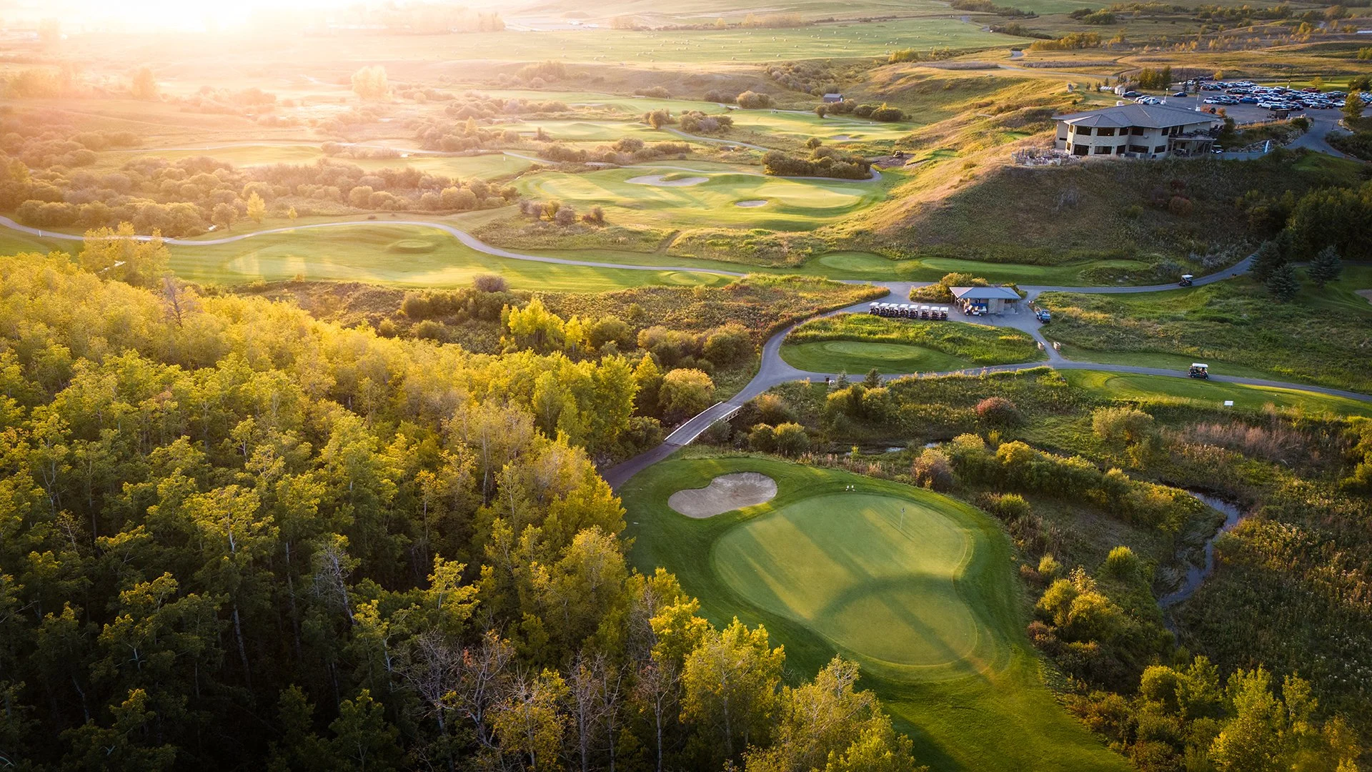 Aerial view of a golf course during sunset with green fairways, sand bunkers, surrounded by trees, with a building and parking lot in the background.