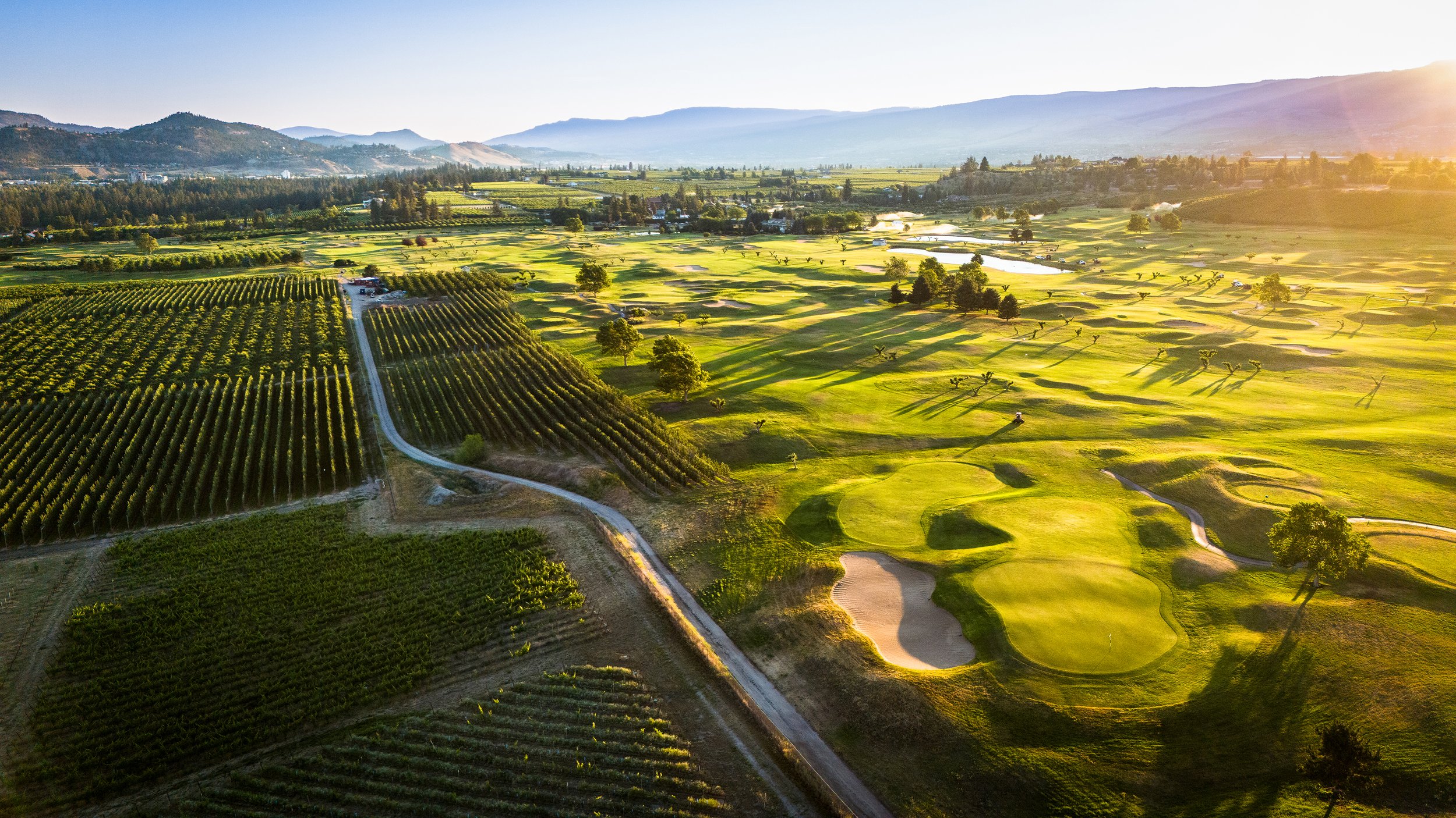 Aerial view of a golf course with sand traps, trees, and surrounding fields, with mountains in the background and sunlight casting long shadows.
