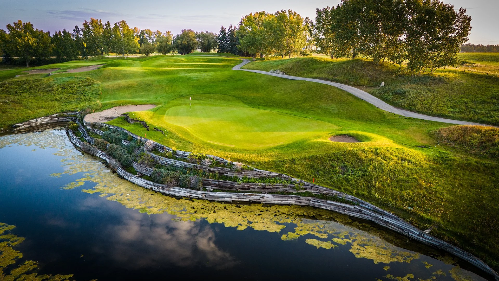 A golf course with a green, sand trap, and water hazard surrounded by trees and lush grass.