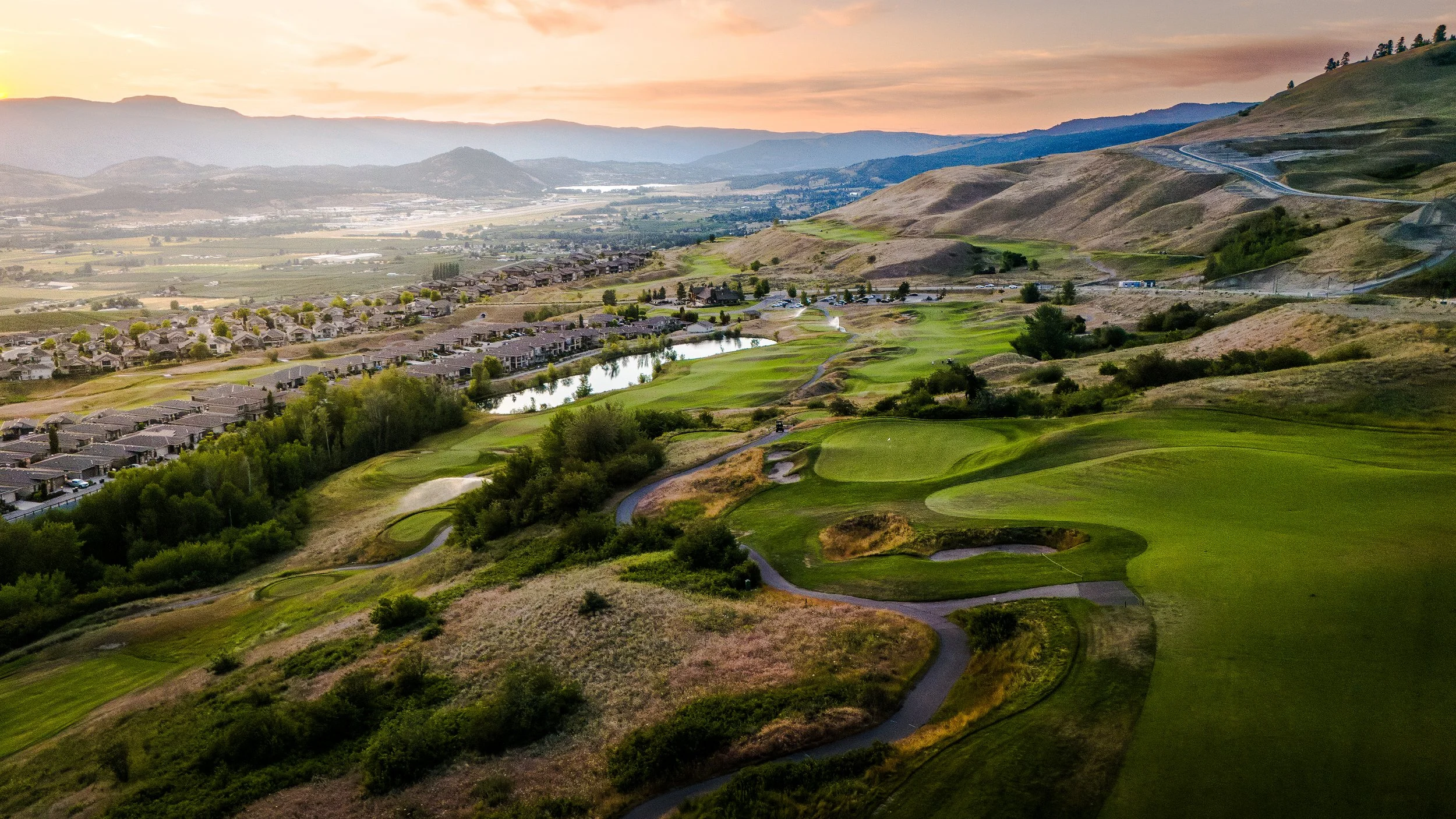 A scenic view of a golf course with lush green fairways, sand traps, and water hazards, situated in a hilly area with surrounding houses, trees, and mountains in the background during sunset.