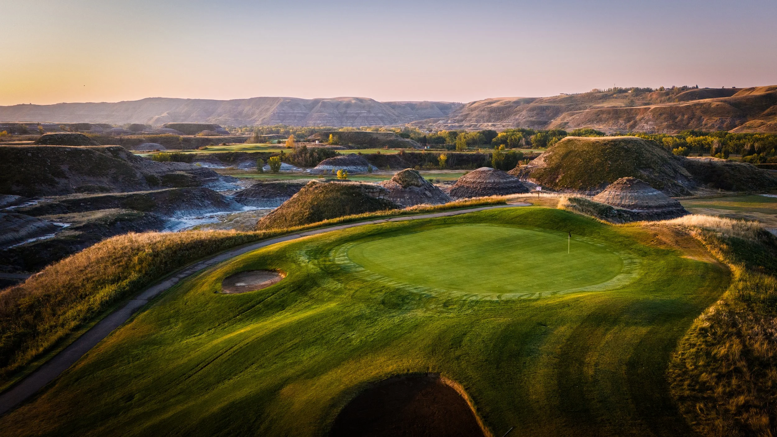A golf course with a green and flag in the foreground, rolling hills and trees in the background at sunset.