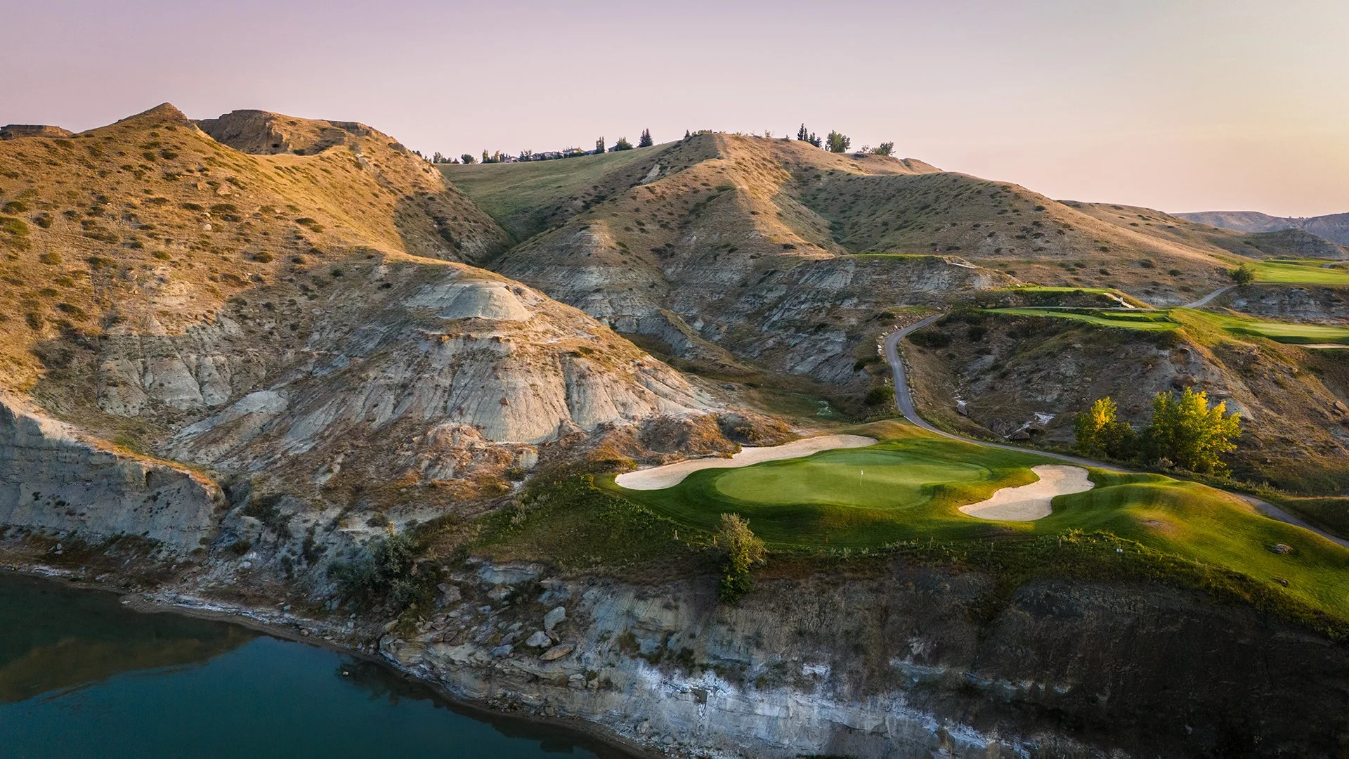 A scenic view of a golf course situated on a hillside with rocky terrain and sparse vegetation, along with a body of water at the base of the hill.