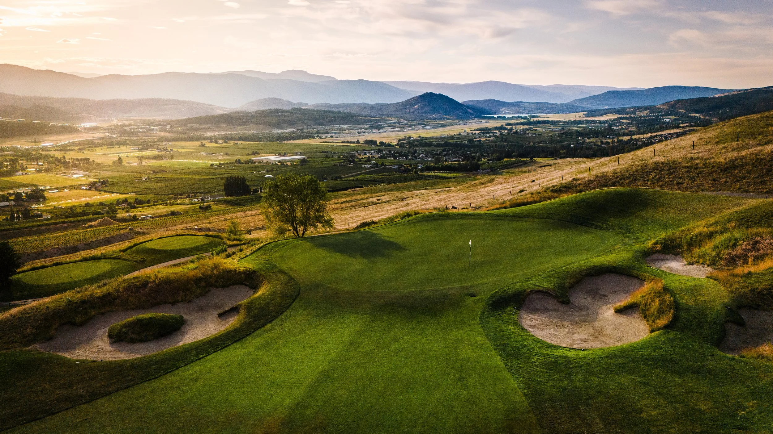 A scenic view of a golf course with green fairways and sand bunkers, set against a backdrop of rolling hills and mountains at sunset.