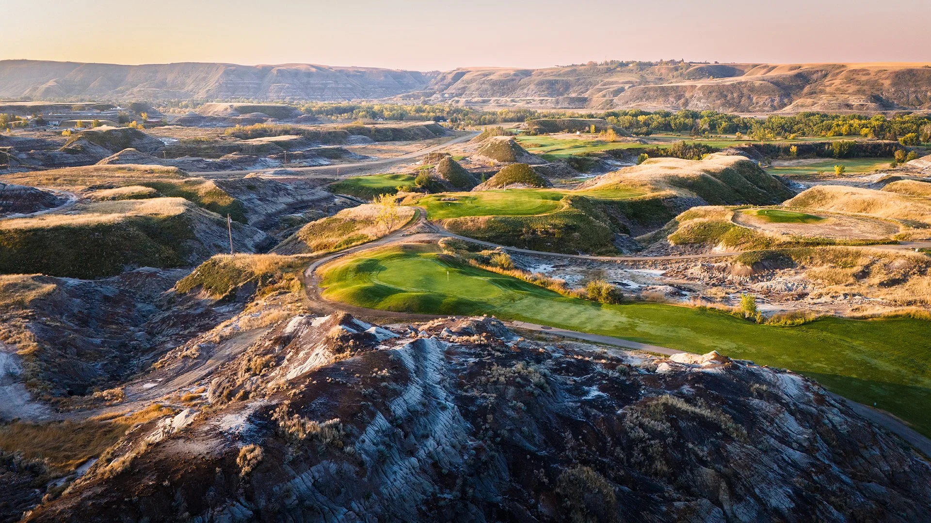 Aerial view of a rugged golf course with green fairways and putting greens, surrounded by dry, rocky hills and sparse vegetation, with a distant landscape under a soft, pinkish sky.