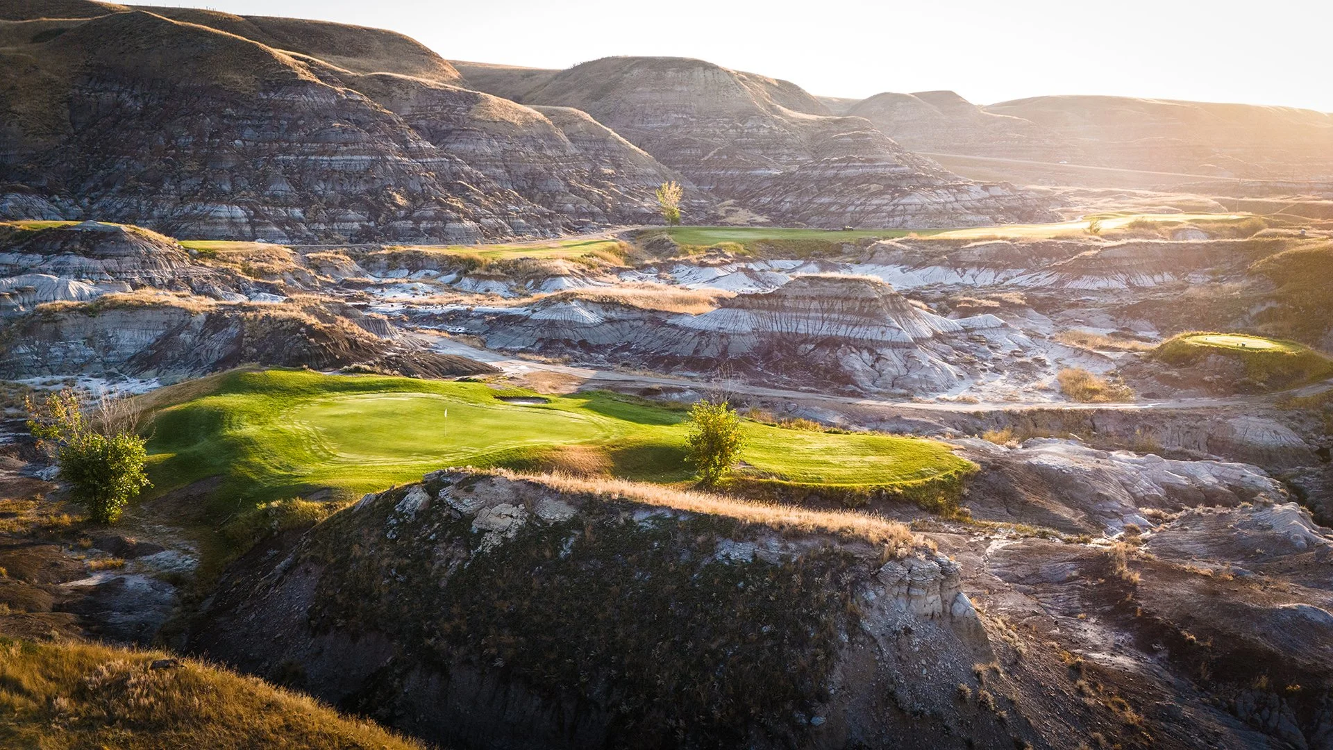 A scenic landscape featuring a golf course with green fairways and small trees, set amid rugged, arid hills and rocky terrain, illuminated by bright sunlight.
