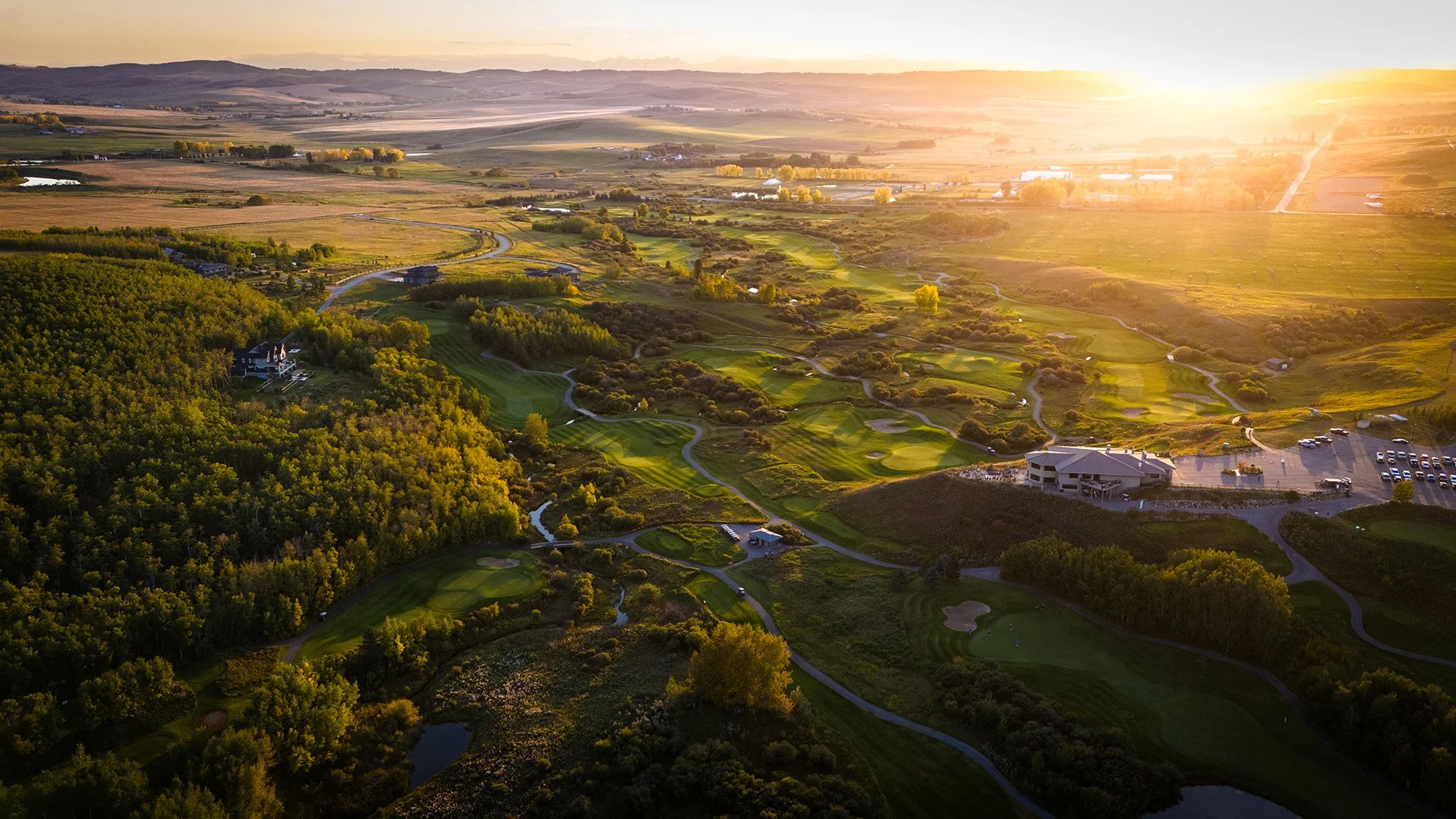 Aerial view of a golf course at sunset with green fairways, sand traps, water hazards, surrounded by trees and nearby buildings and parking lot.