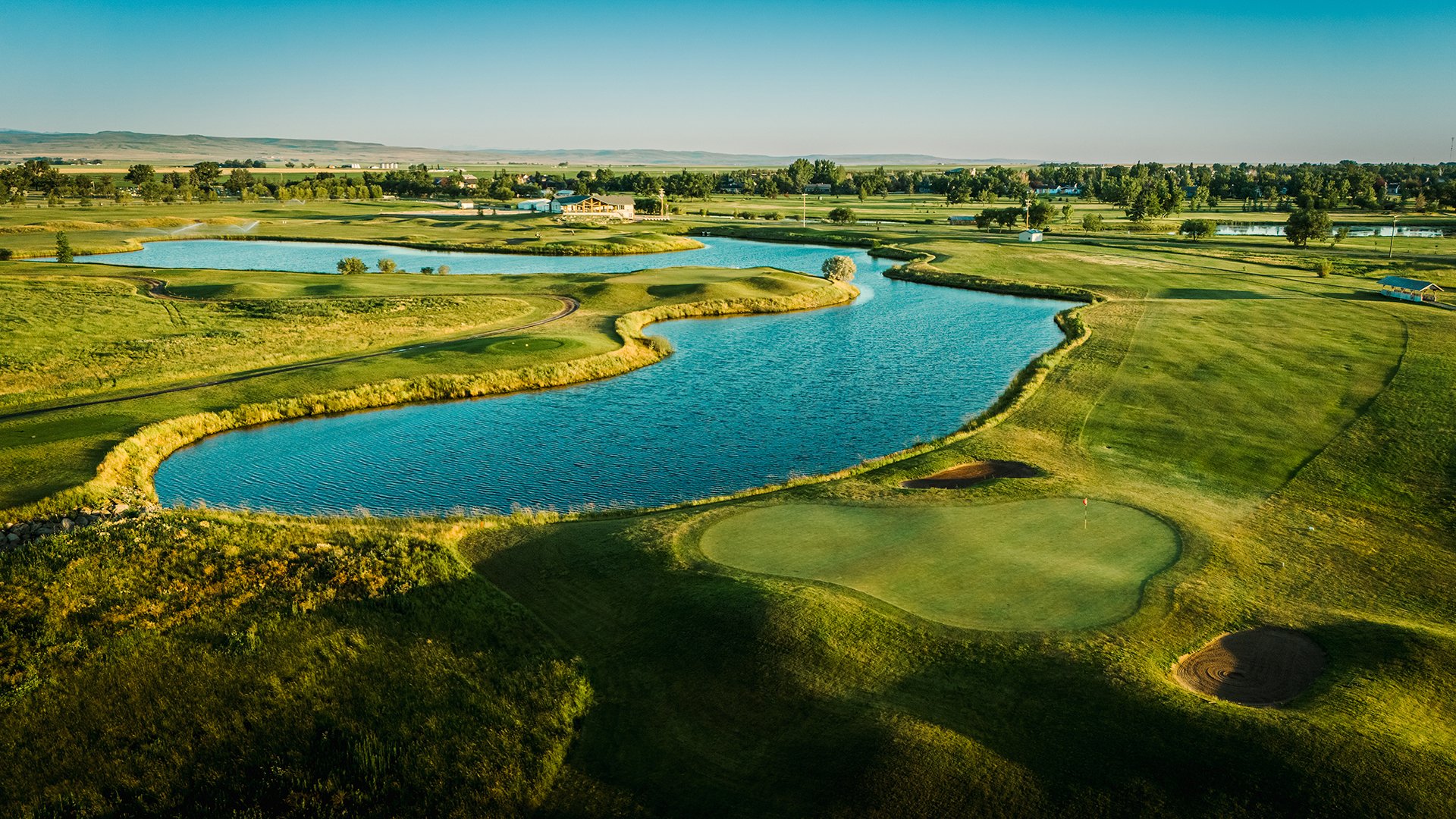 Aerial view of a golf course with water hazards and green fairways, surrounded by trees and open fields under a clear blue sky.