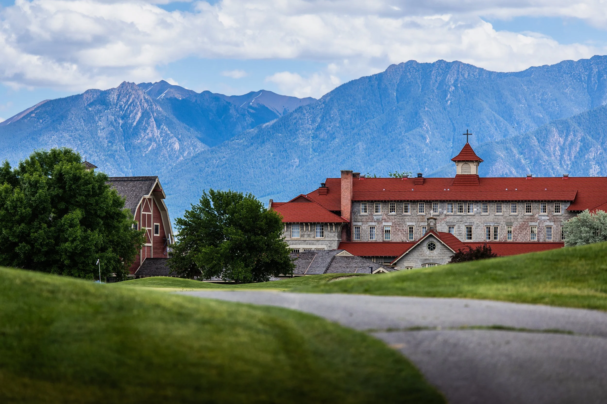 A scenic view of a church with a red roof and cross, surrounded by trees, with mountains and blue sky in the background.
