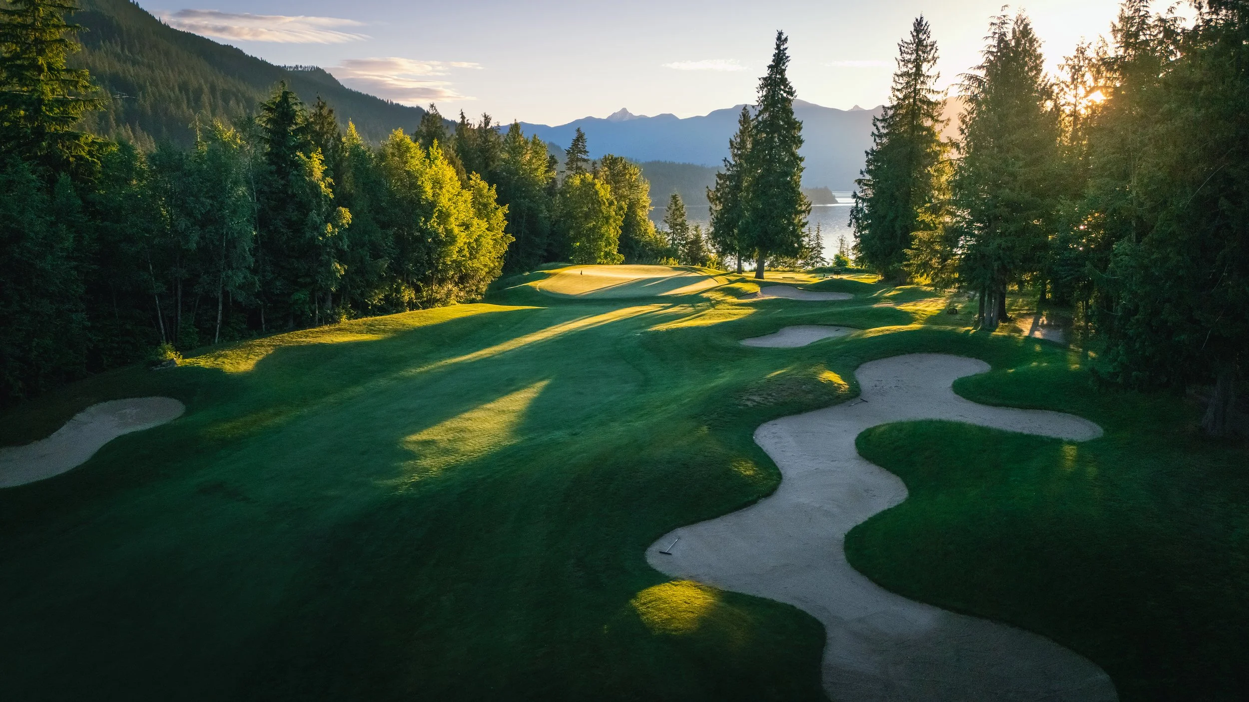 Sunset over a lush green golf course surrounded by trees and mountains in the distance.