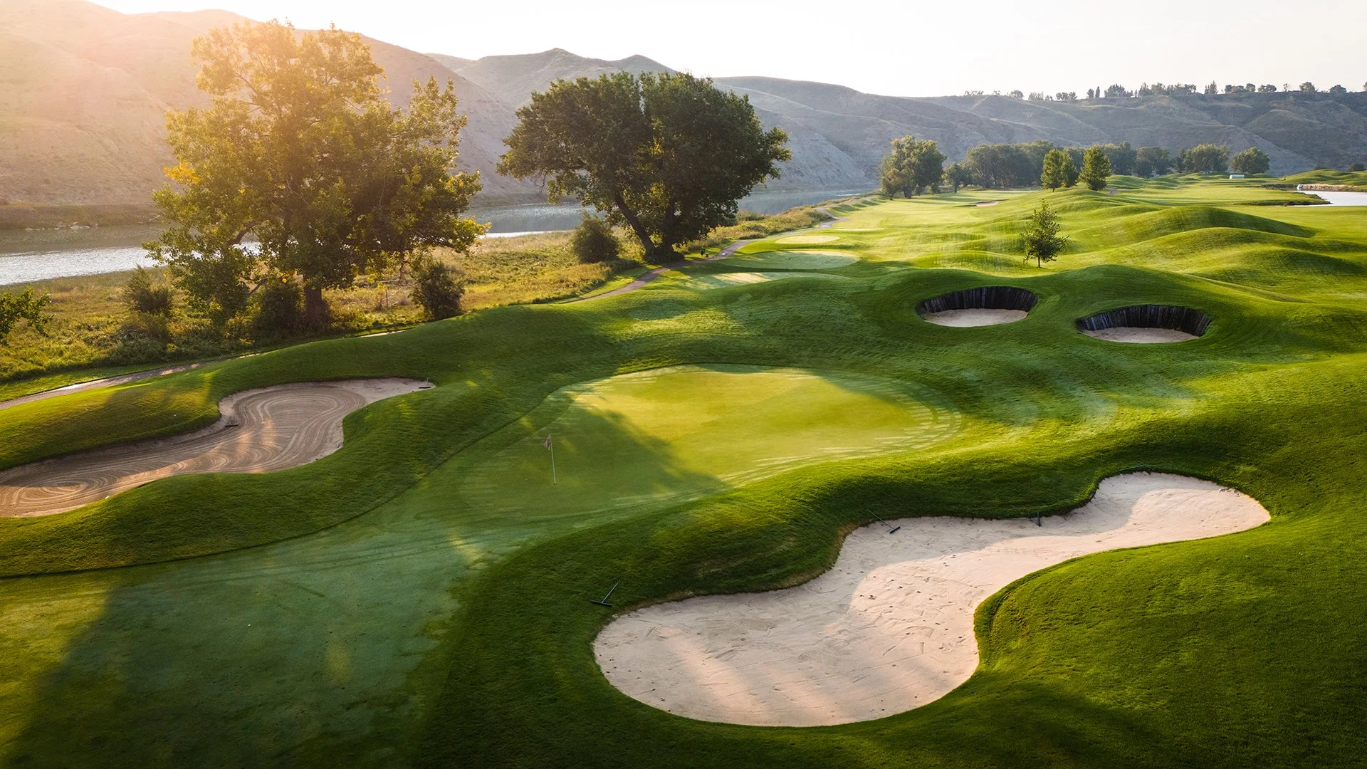 A scenic golf course featuring sand bunkers, a putting green with a flag, lush grass, trees, water, and distant mountains under a sunny sky.