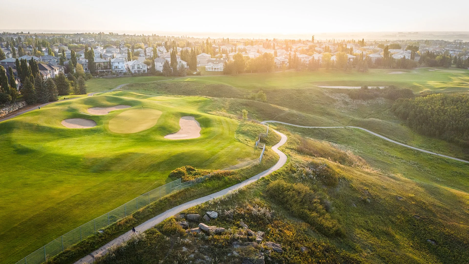 Aerial view of a golf course at sunset with houses in the background and a winding path in the foreground.