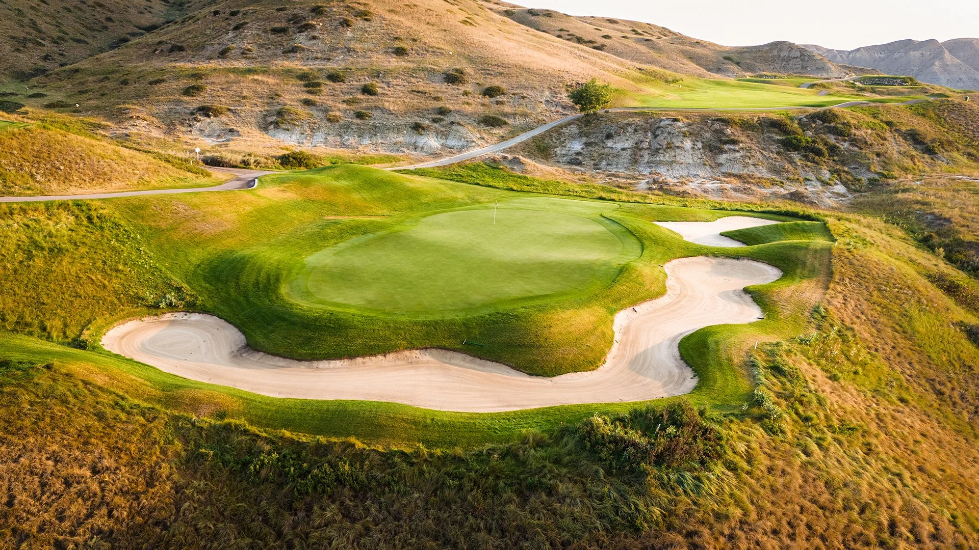An aerial view of a golf course with green fairways, sand traps, and a flag marking the hole, surrounded by hills and sparse vegetation.