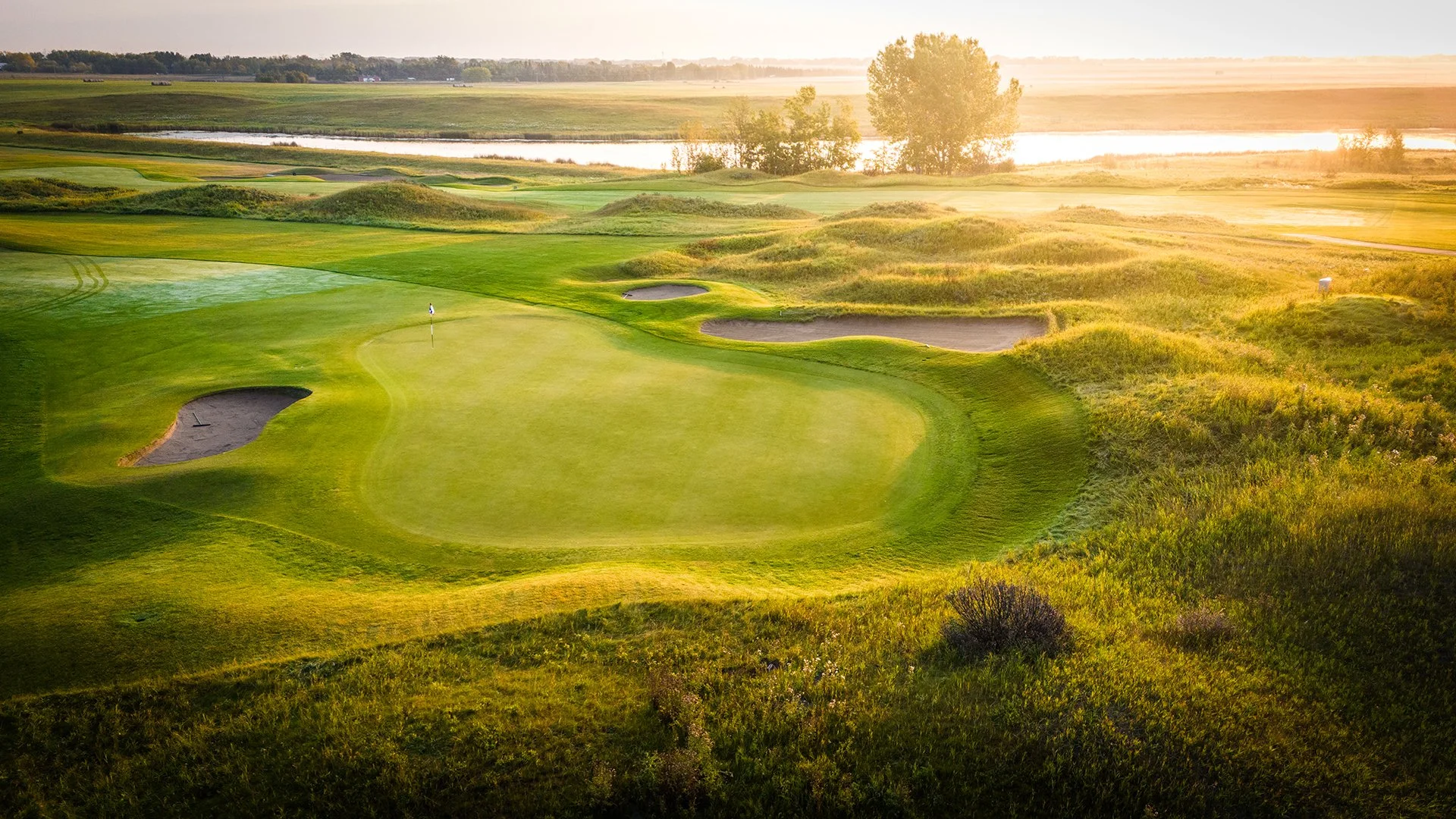 Sunset over a golf course with green, sand bunkers, and water hazards, surrounded by lush grass and trees.