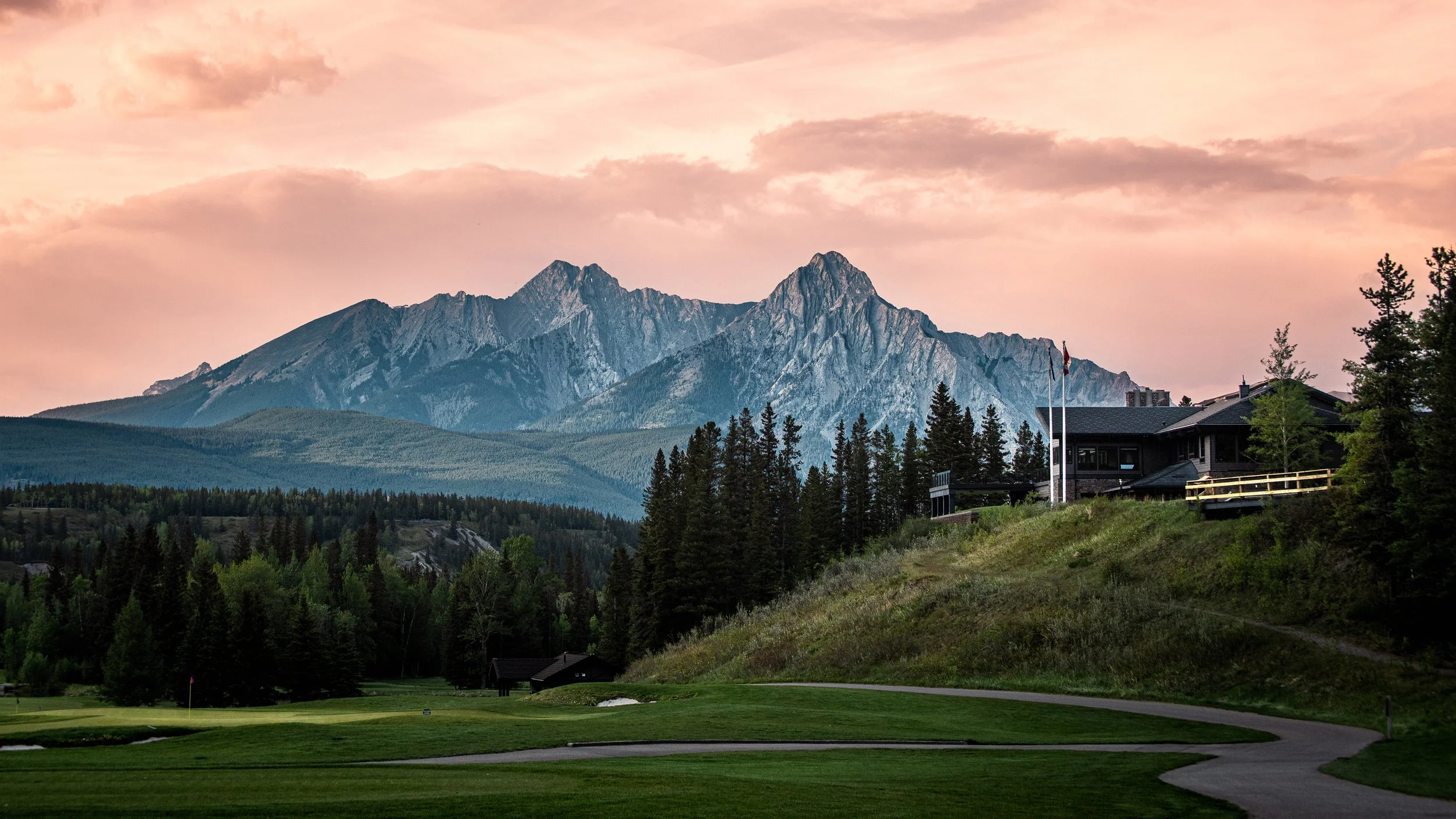 A scenic view of a mountain range at sunset with a pink sky, forested hills, and a house with a deck among trees.