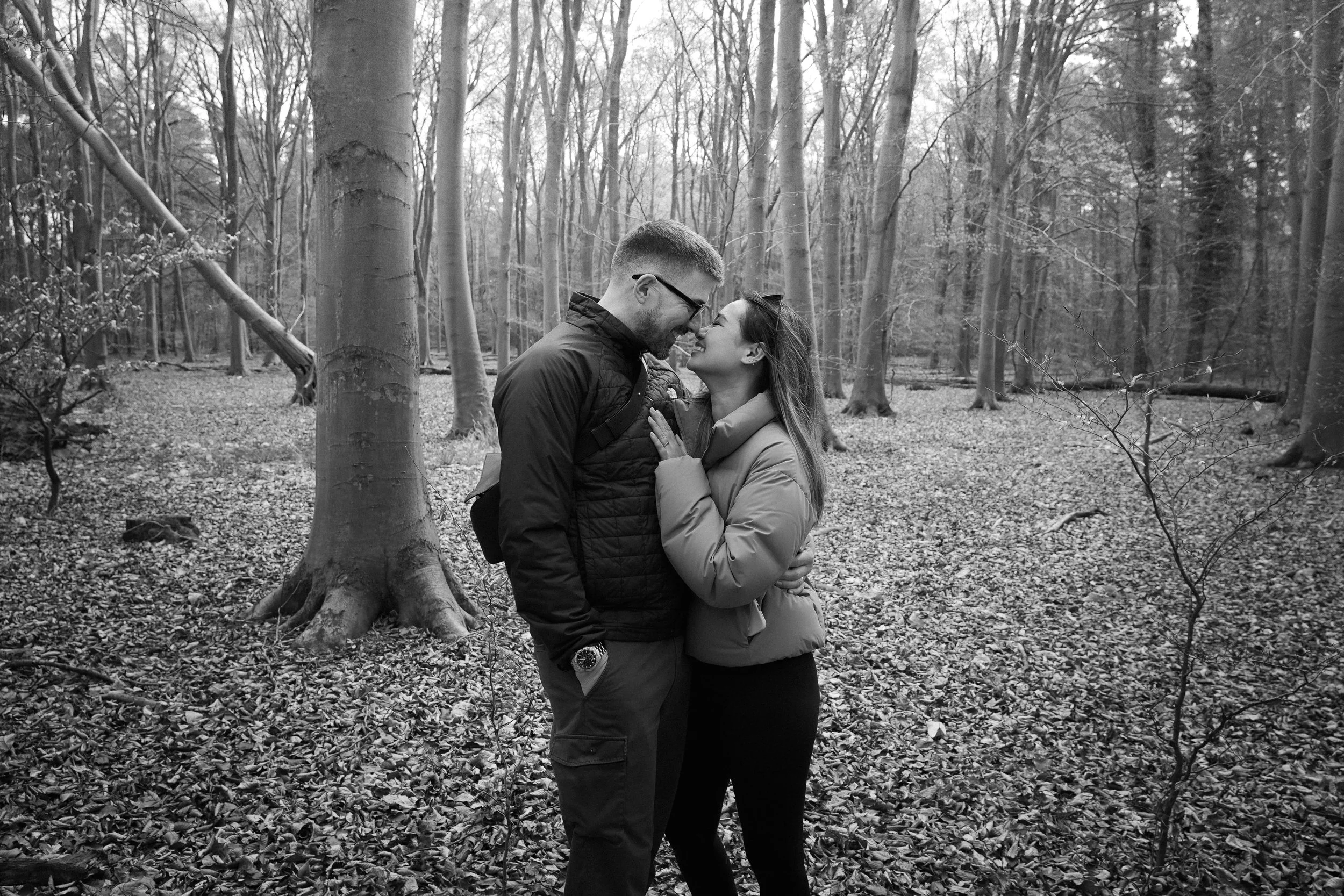 A happy couple in a forest, standing close and touching foreheads. The man is wearing glasses and a jacket, and the woman is wearing a puffy coat. They are smiling with their noses close together.